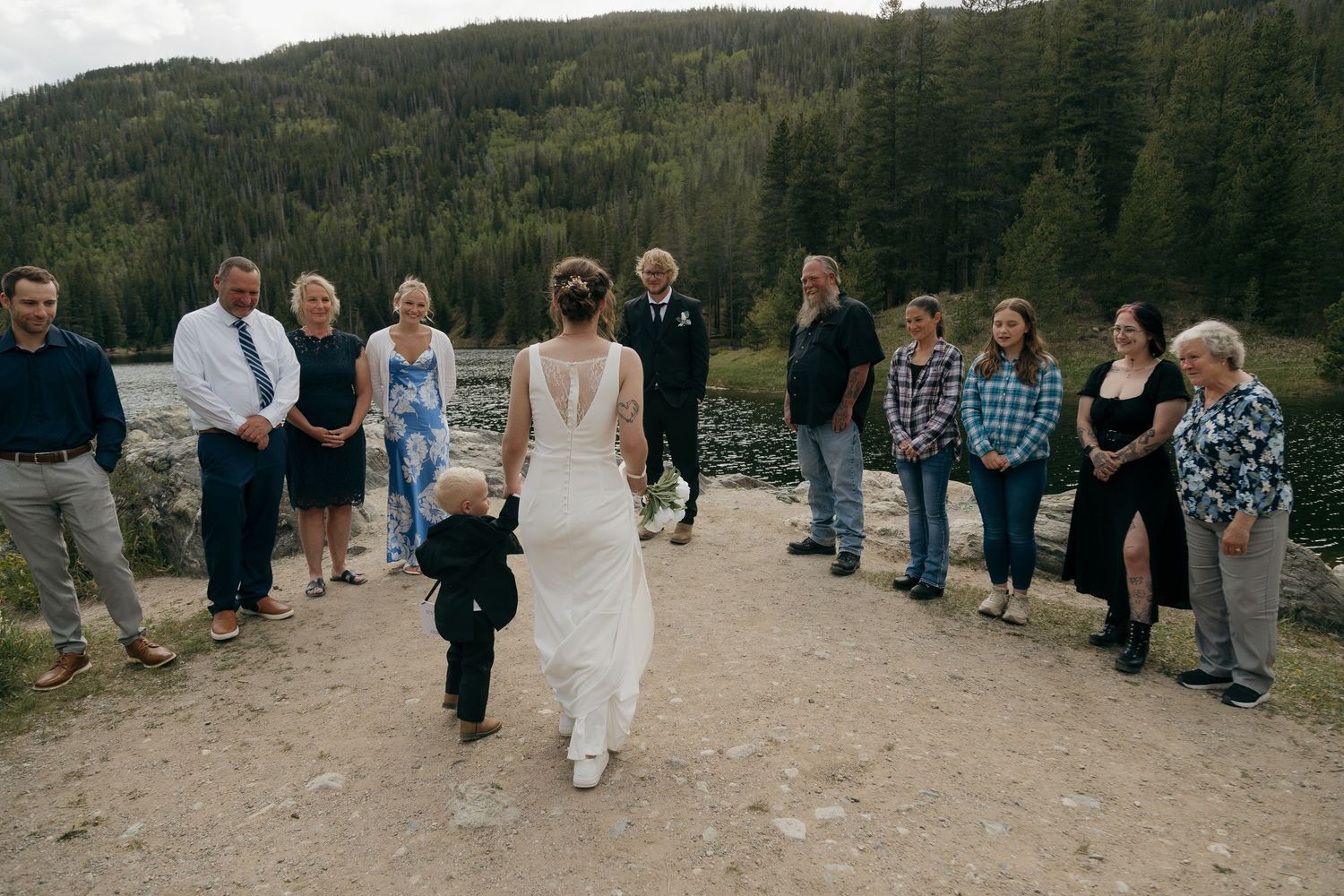 Bride walks with young boy in tuxedo toward groom at outdoor wedding ceremony with mountain forest backdrop.
