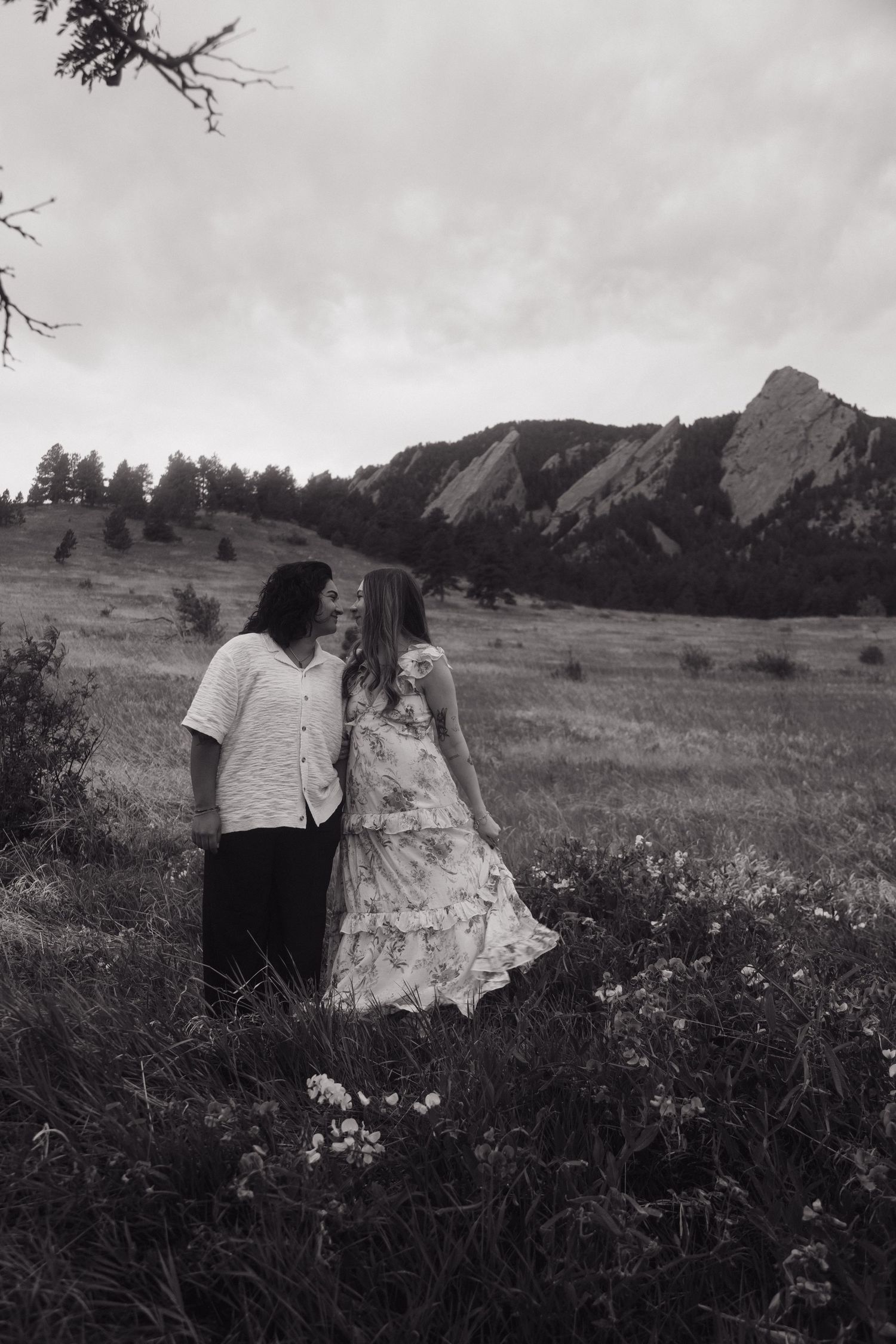 Black and white photo of a couple holding hands in a wildflower meadow with the Flatirons mountains in the background.