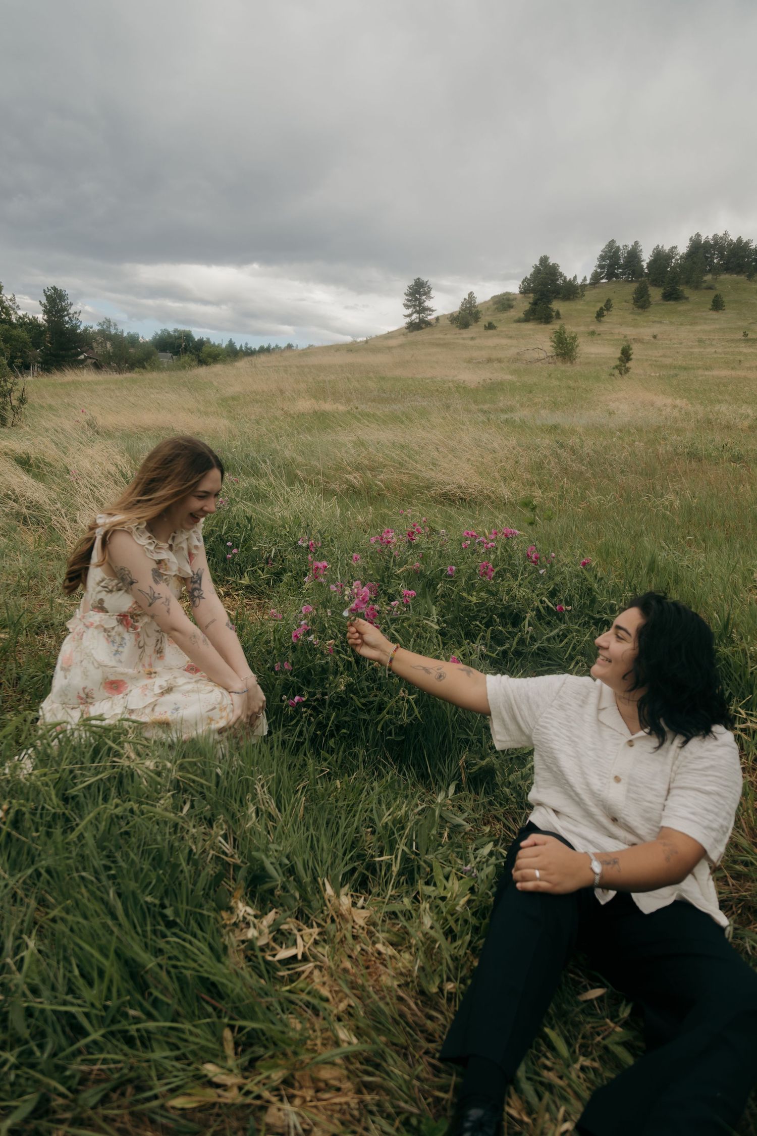 A couple sits in a wildflower meadow as one offers a pink flower to the other on an overcast day.