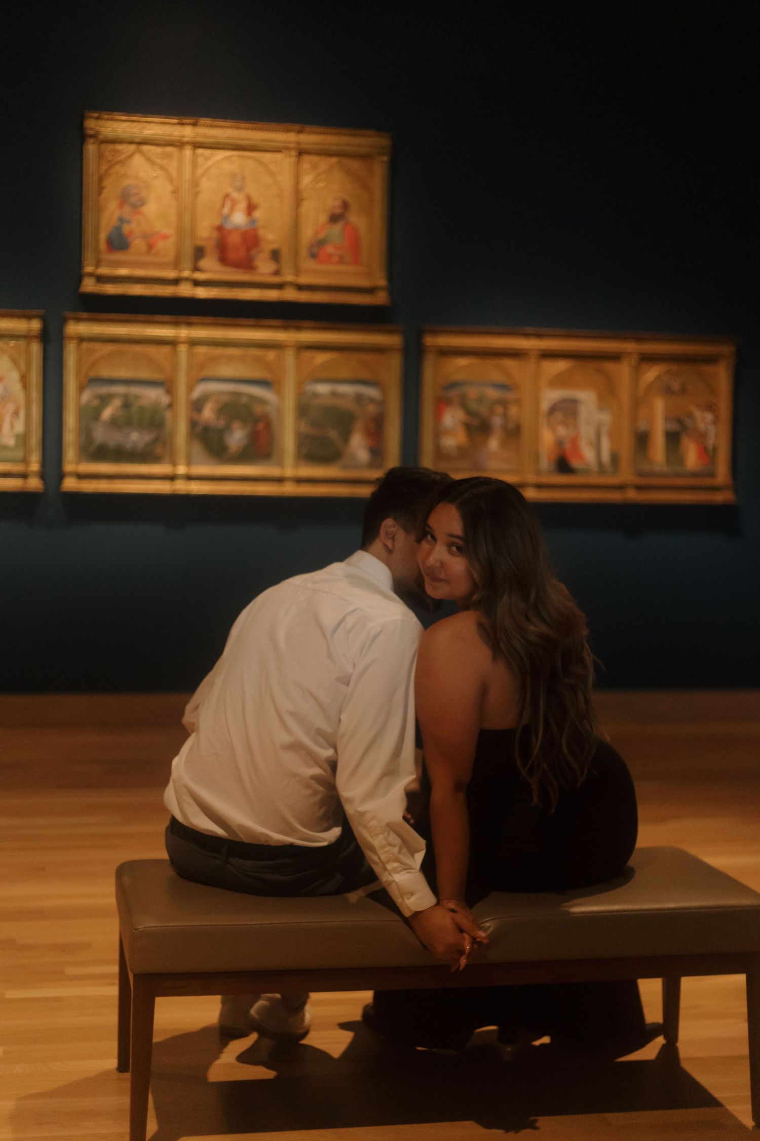 Couple holding hands on a museum bench admiring Renaissance paintings on a dark wall.