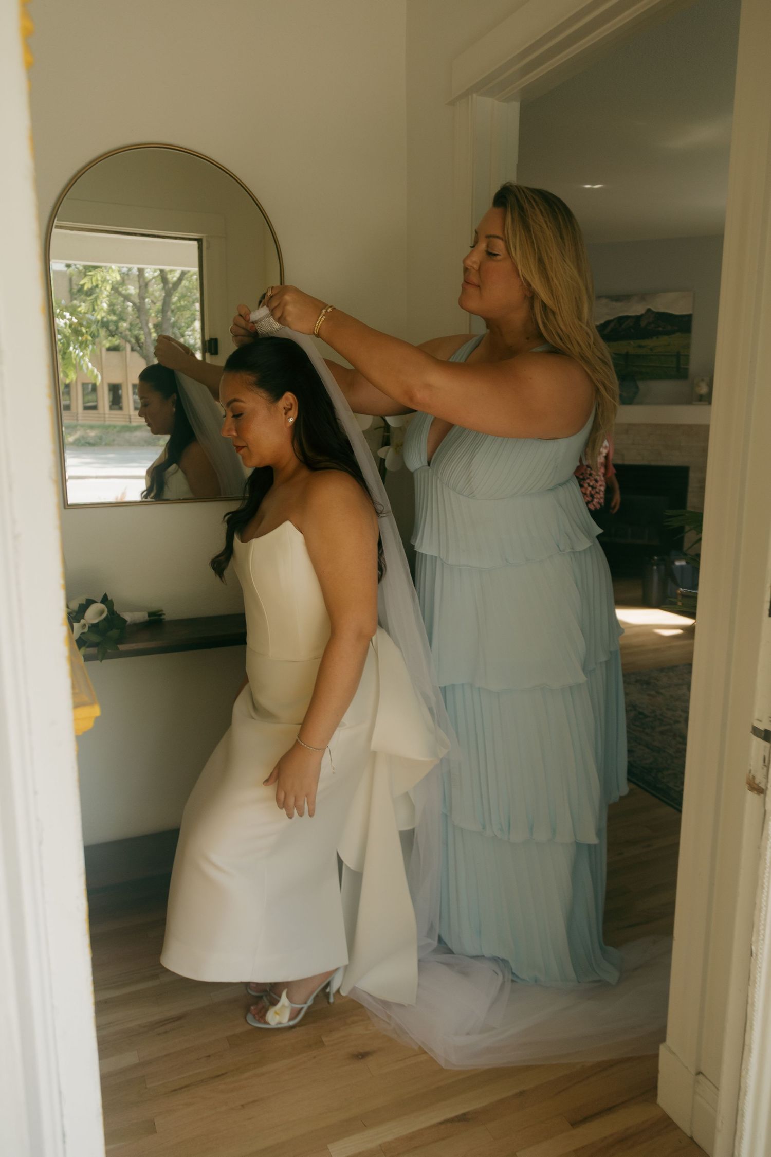 Bridesmaid in sage green dress helps bride with veil in front of mirror during wedding preparations.