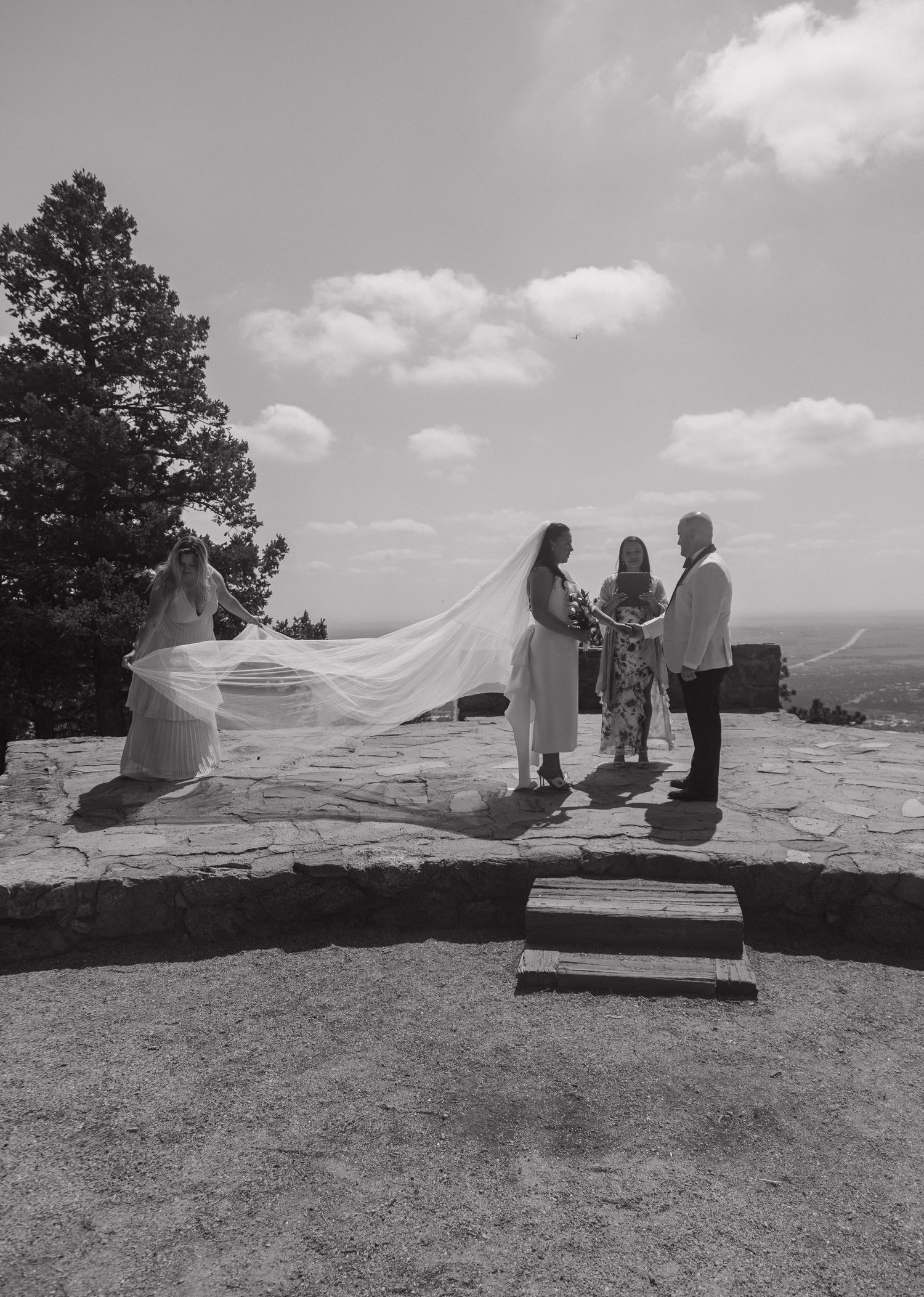 Black and white photo of a bride and groom exchanging vows outdoors on a rocky hilltop with a flowing veil in the wind.