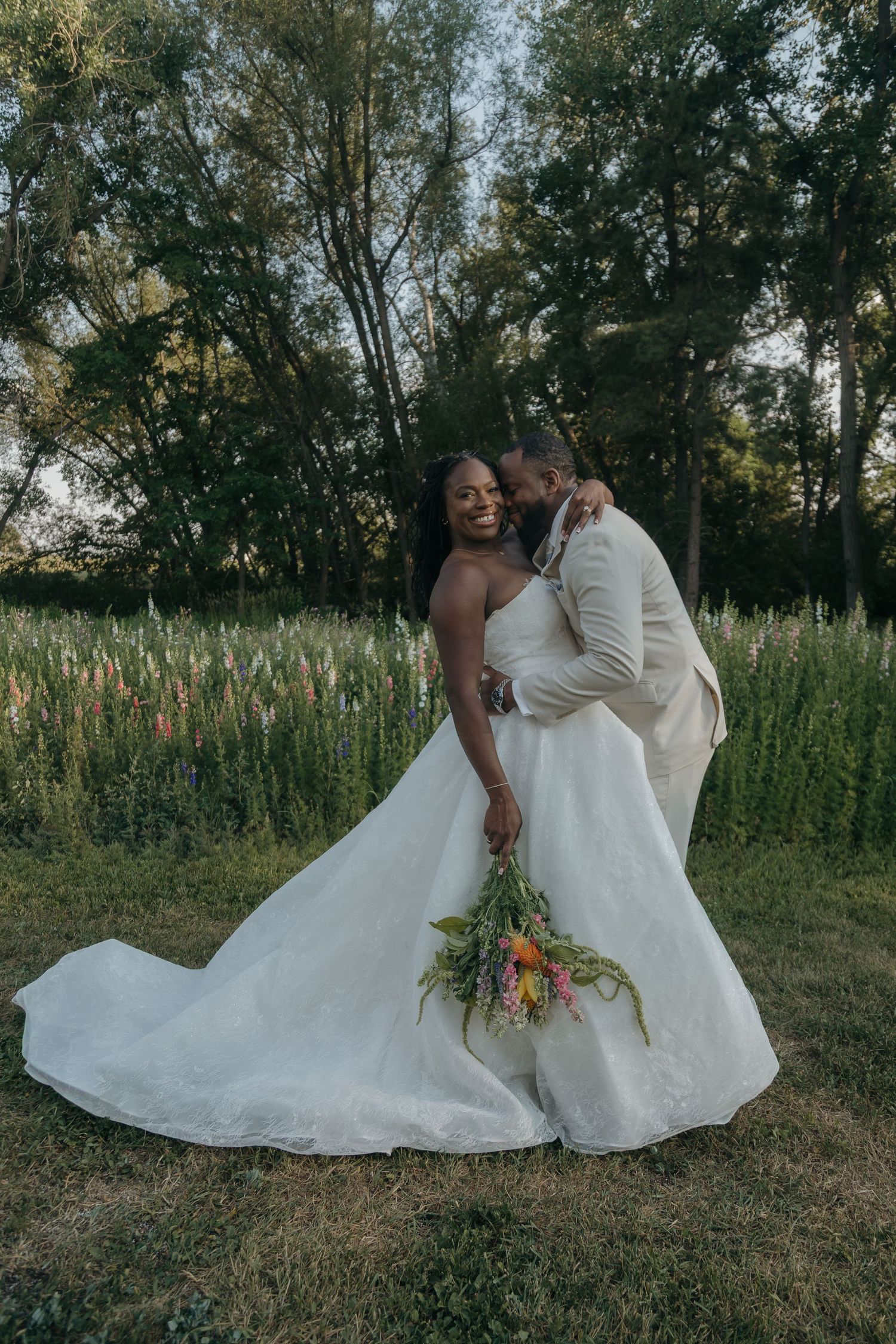 Bride in white gown laughs as groom in cream suit kisses her cheek outdoors, holding colorful wildflower bouquet.