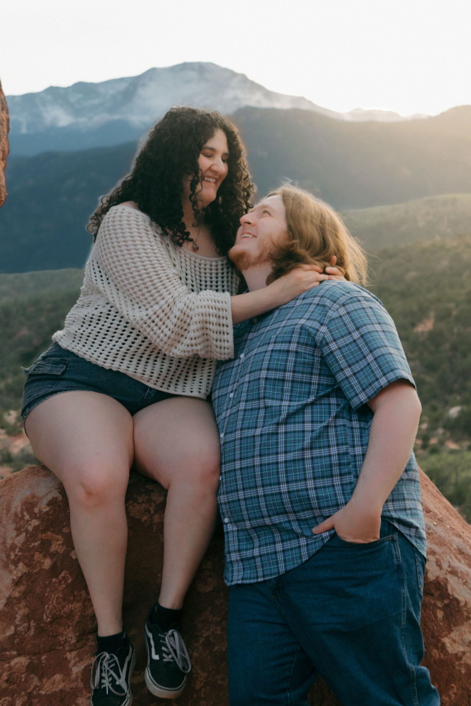 A couple shares laughter and embrace while sitting on red rocks with mountain views.