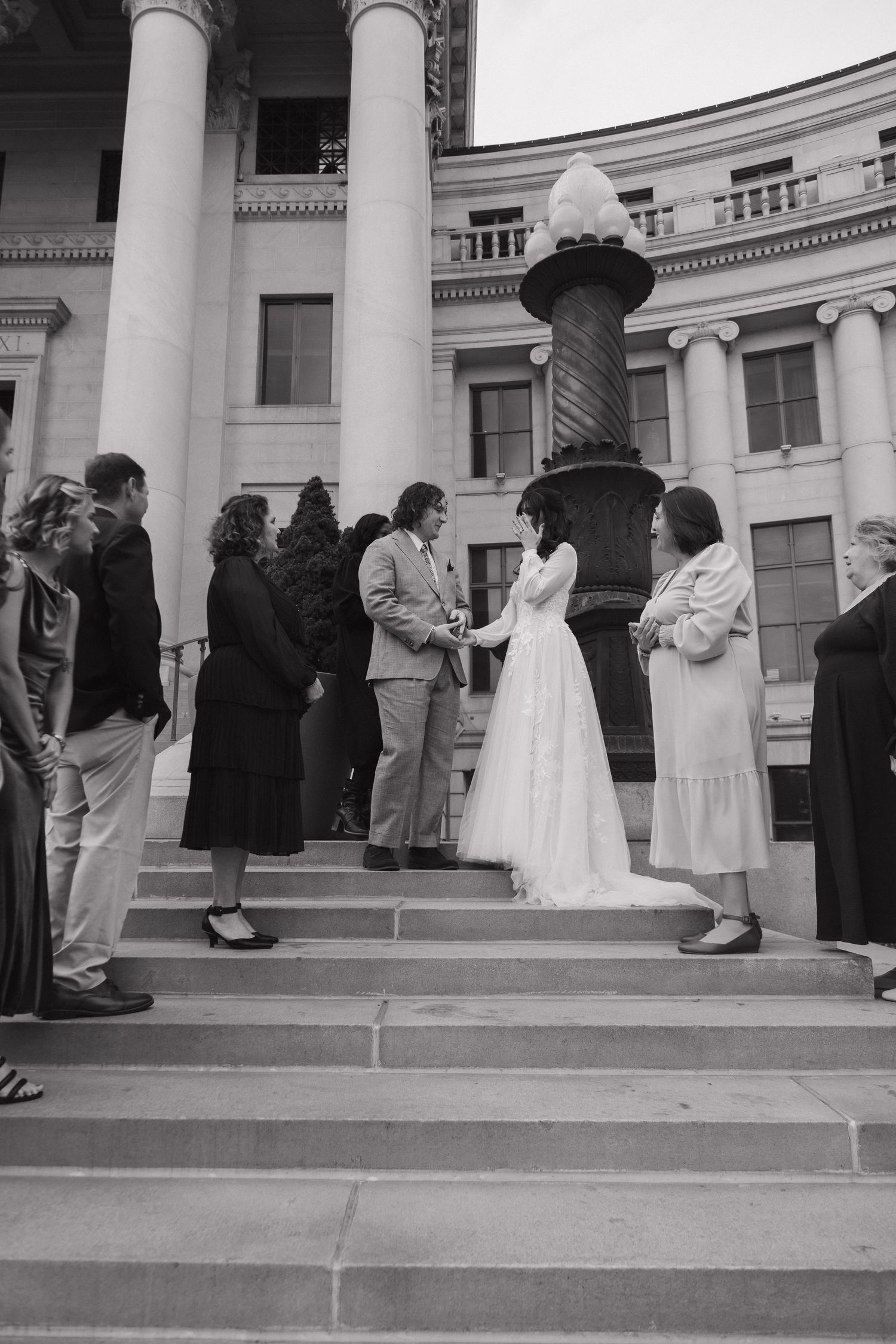 A wedding ceremony takes place on the steps of a grand building with white columns and gathered guests.