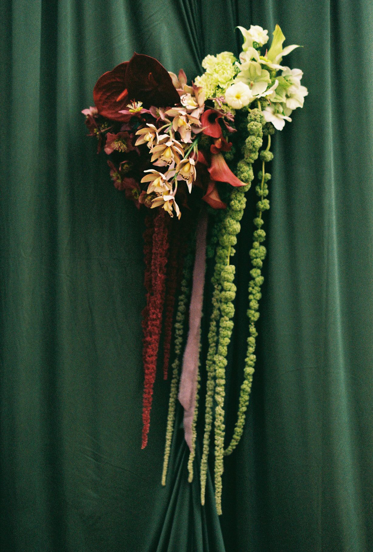 Elegant floral arrangement with cascading burgundy and lime green flowers against a dark emerald green curtain backdrop.