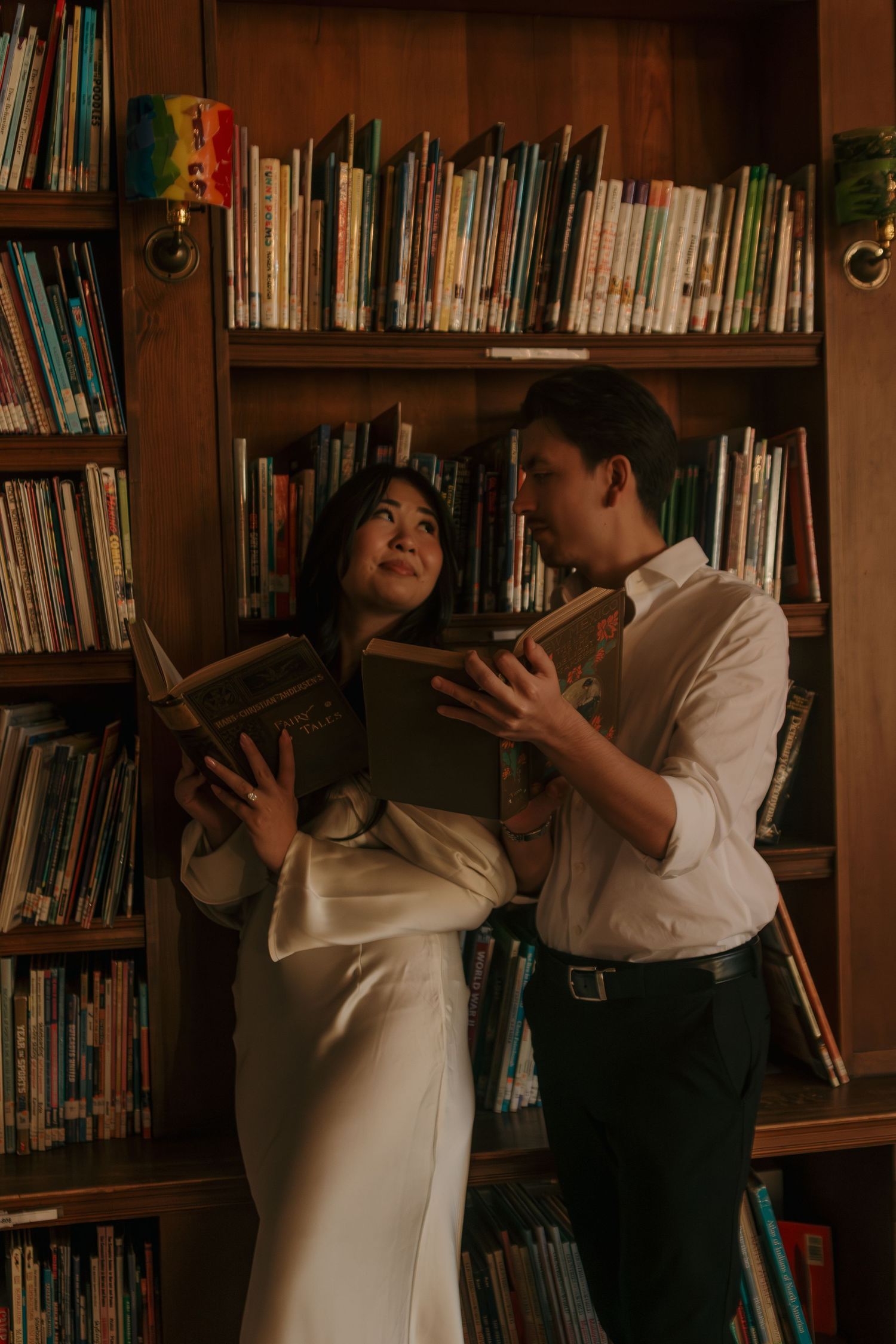 Two people playfully posing with books in a library setting with warm ambient lighting.
