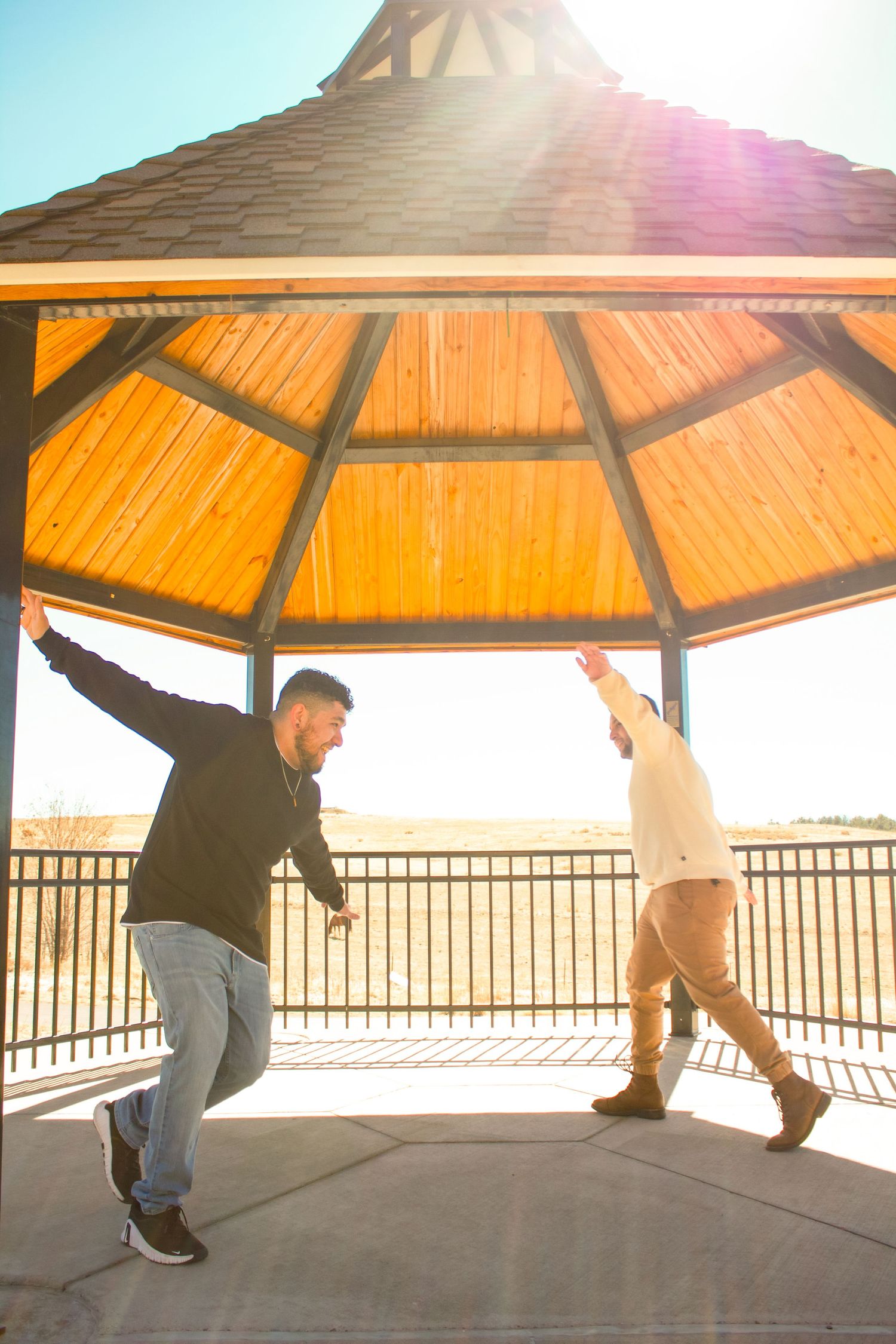 Two people strike playful poses under a wooden gazebo in bright sunlight.