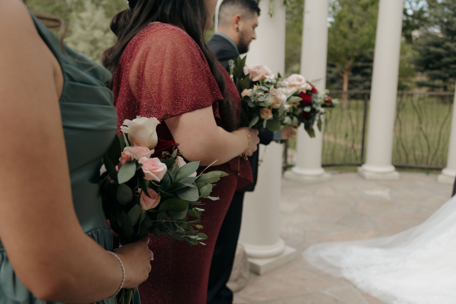 Bridesmaids in burgundy and green gowns holding pink and white floral bouquets during outdoor wedding ceremony.