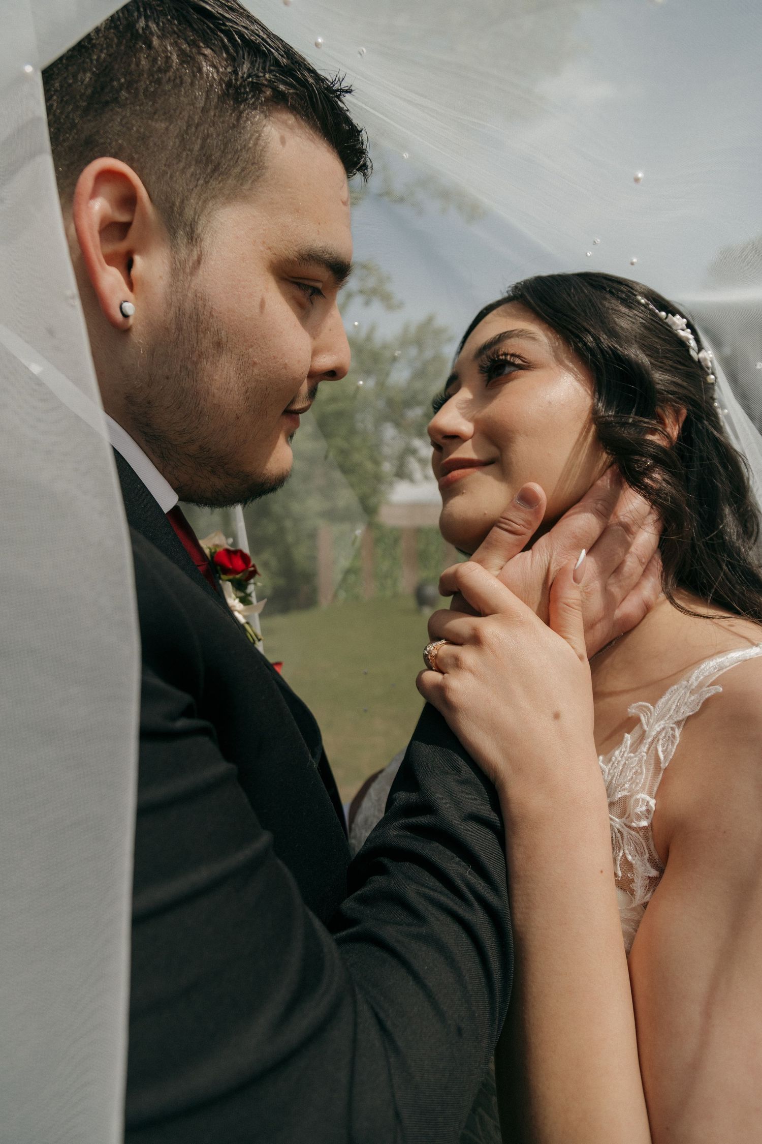 Groom tenderly holding bride's face under wedding veil in an intimate outdoor portrait.