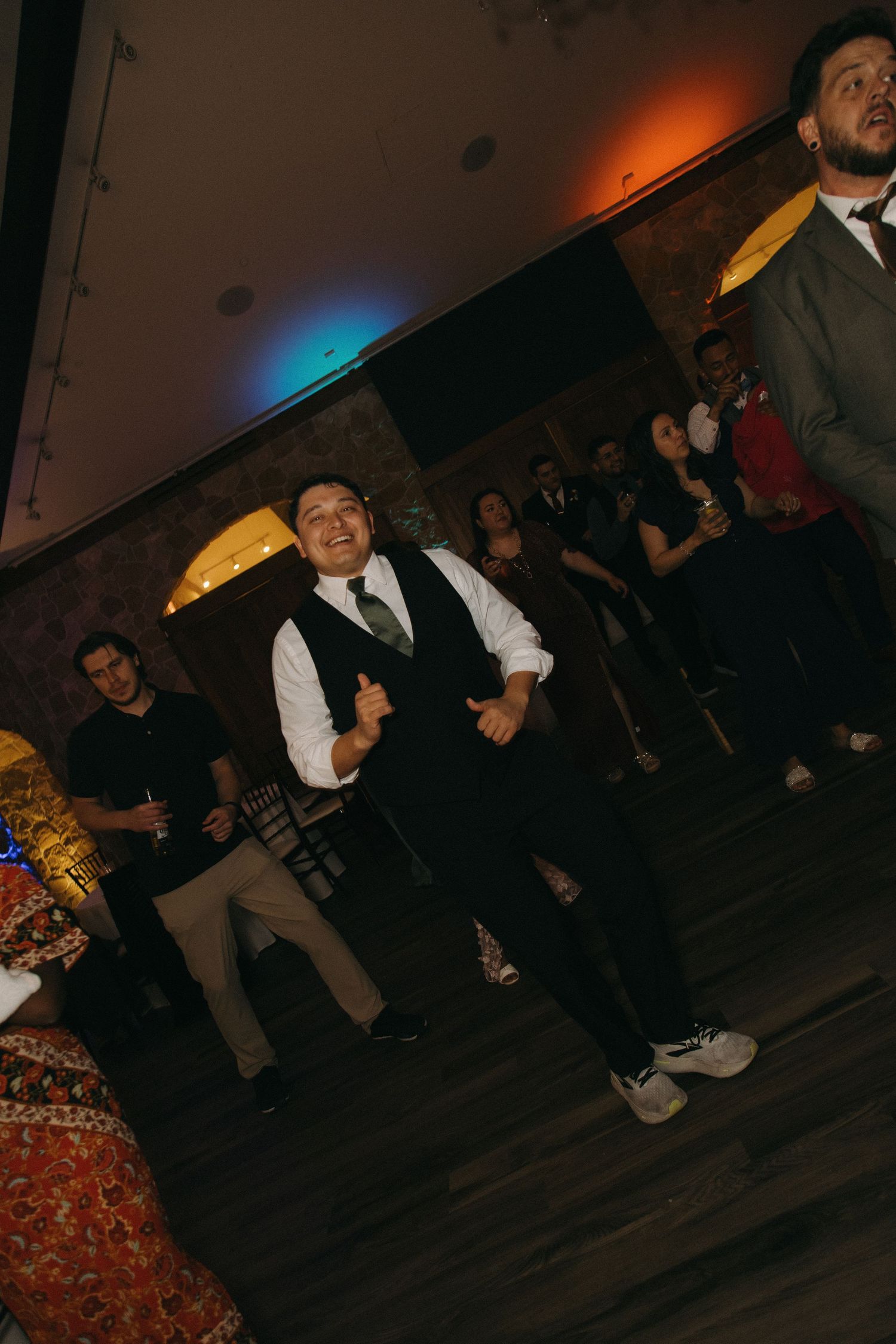 Groomsman in black vest dances joyfully on the dance floor at a lively wedding reception.