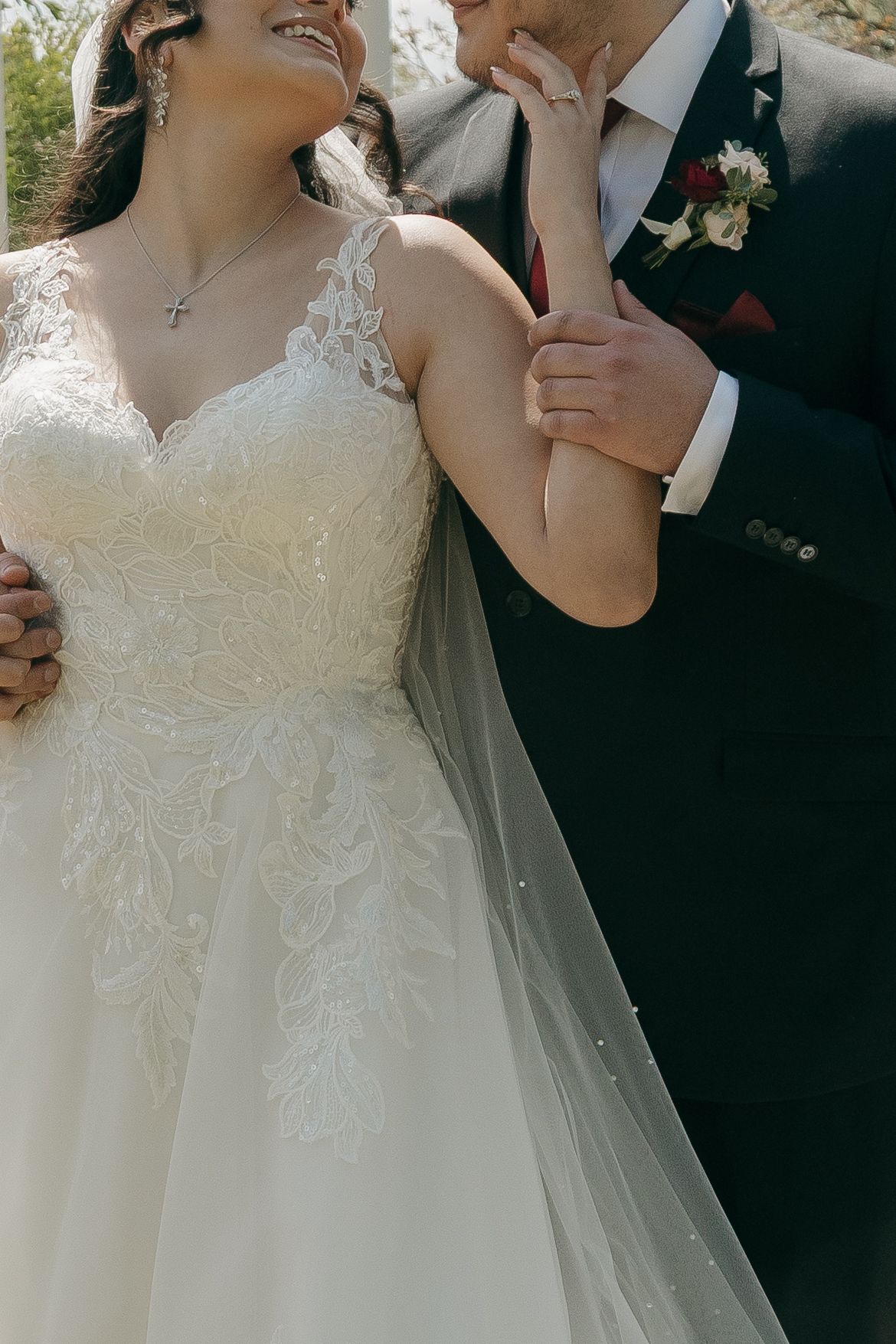 Close-up of a bride in lace wedding gown and groom in navy suit sharing a joyful moment outdoors.
