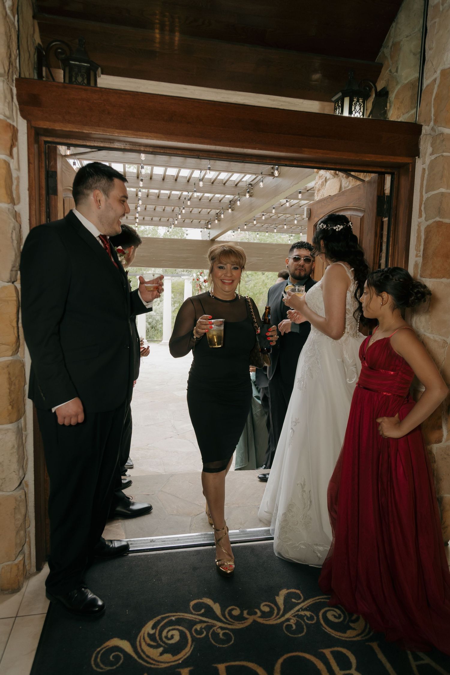 Wedding party gathers at venue entrance with bride in white gown and guests holding drinks celebrating.