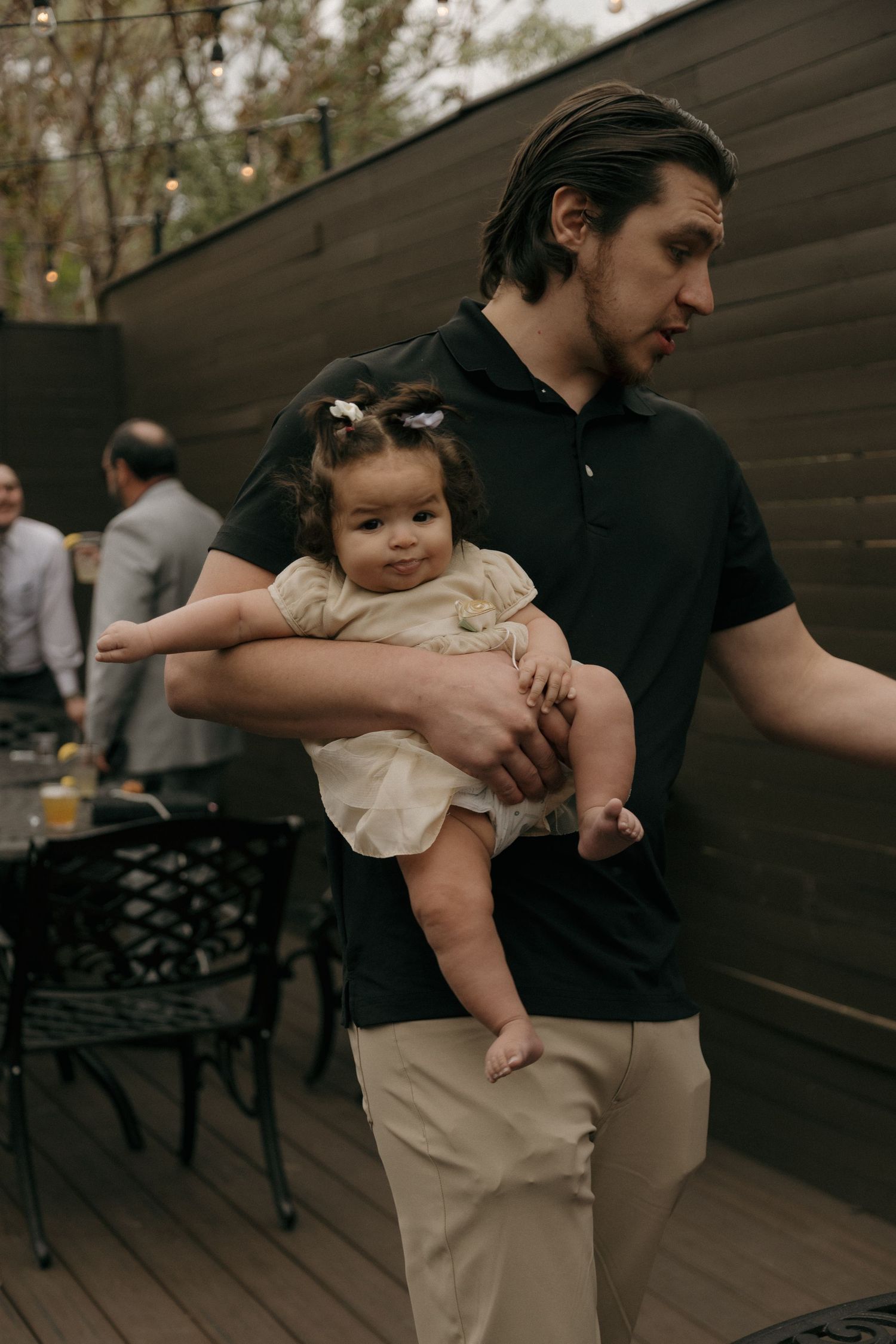 Young man in black polo shirt holding a smiling baby girl in a cream dress at an outdoor wedding reception.