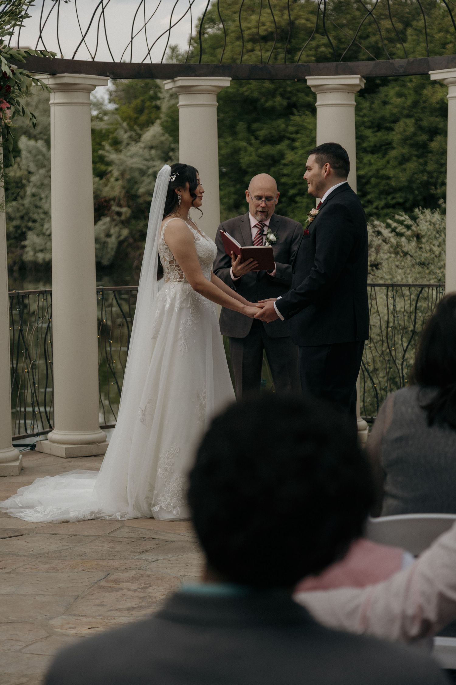 Bride and groom hold hands before officiant at elegant outdoor ceremony beneath white columned pergola.