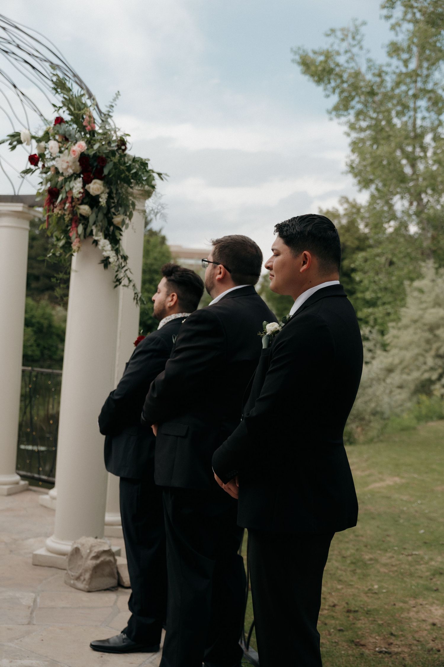 Three groomsmen in black suits stand at outdoor wedding altar decorated with floral arch and white columns.