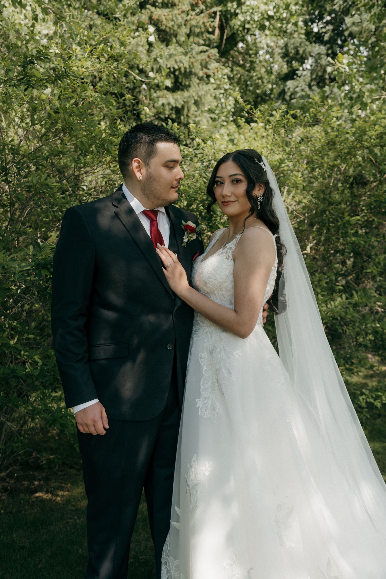 Bride in lace gown with veil and groom in black suit with red tie posing in green garden.