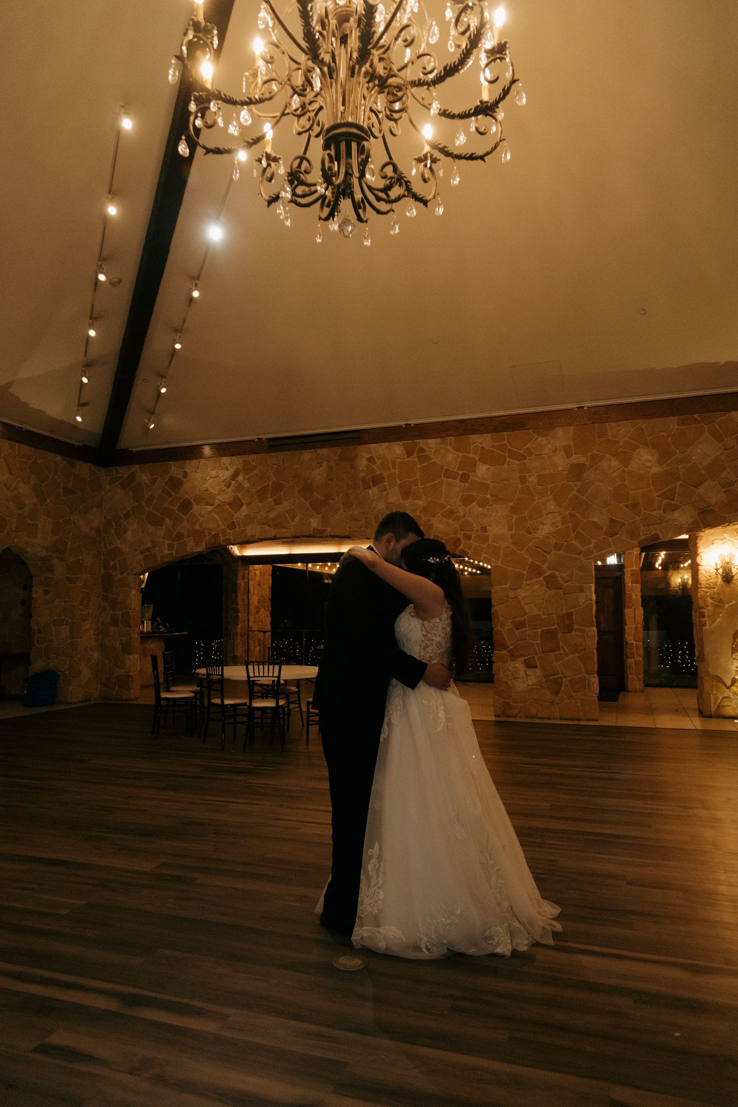 Bride and groom share their first dance beneath an elegant chandelier in a warmly lit rustic wedding venue.