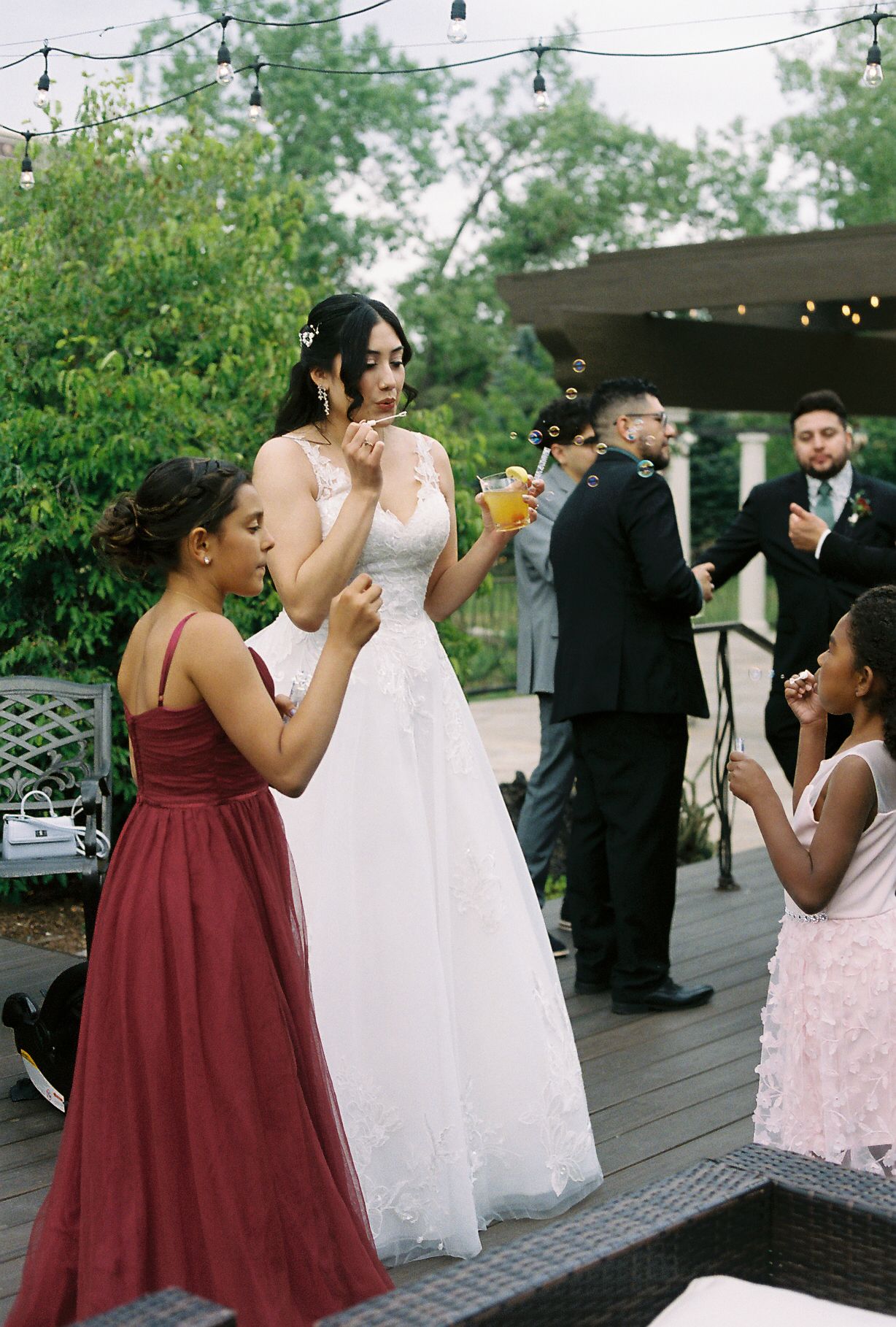 A bride in a white lace gown blows bubbles with young girls in burgundy and pink dresses at a wedding reception.