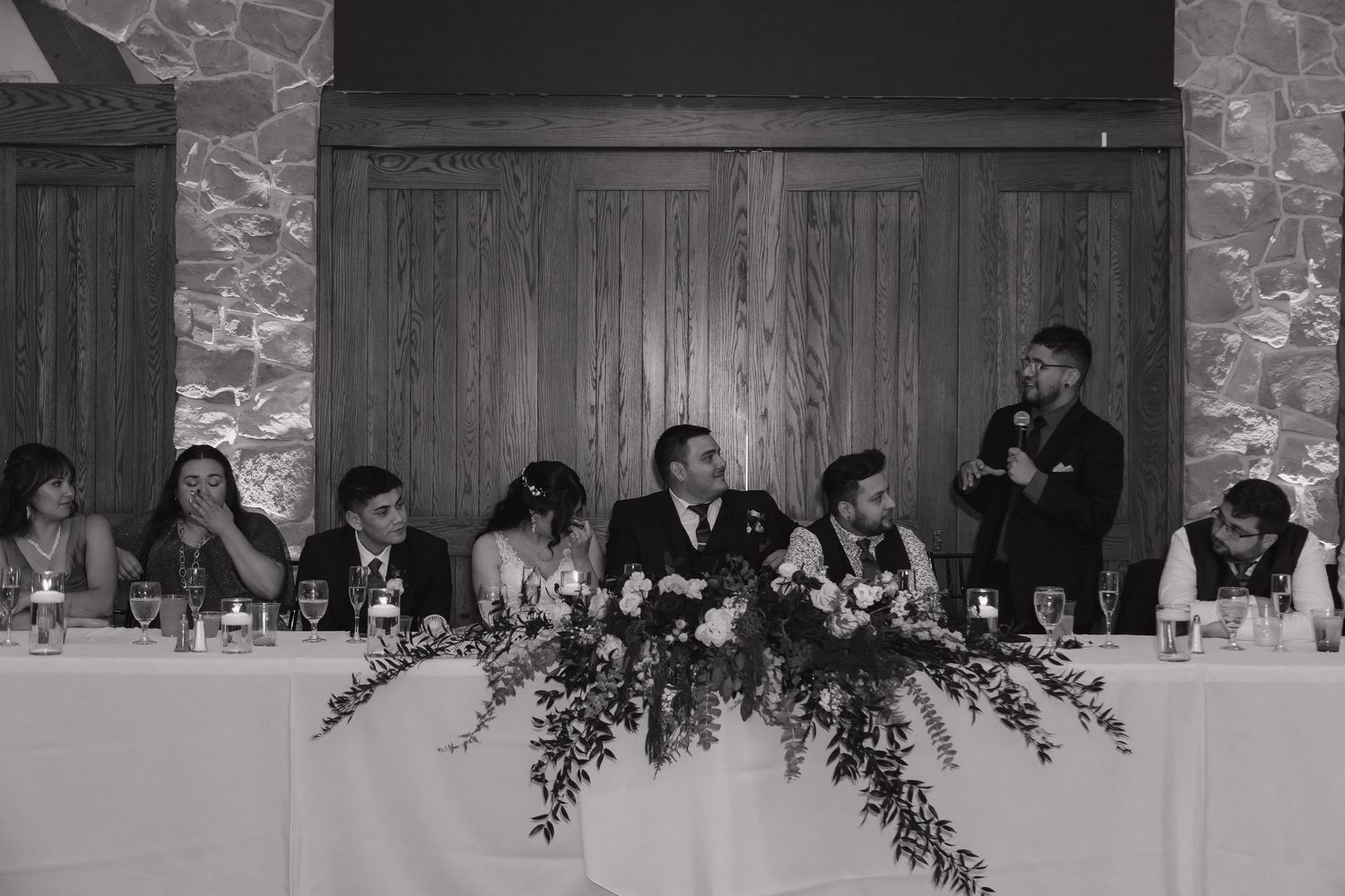 Black and white photo of wedding party seated at decorated head table during reception.