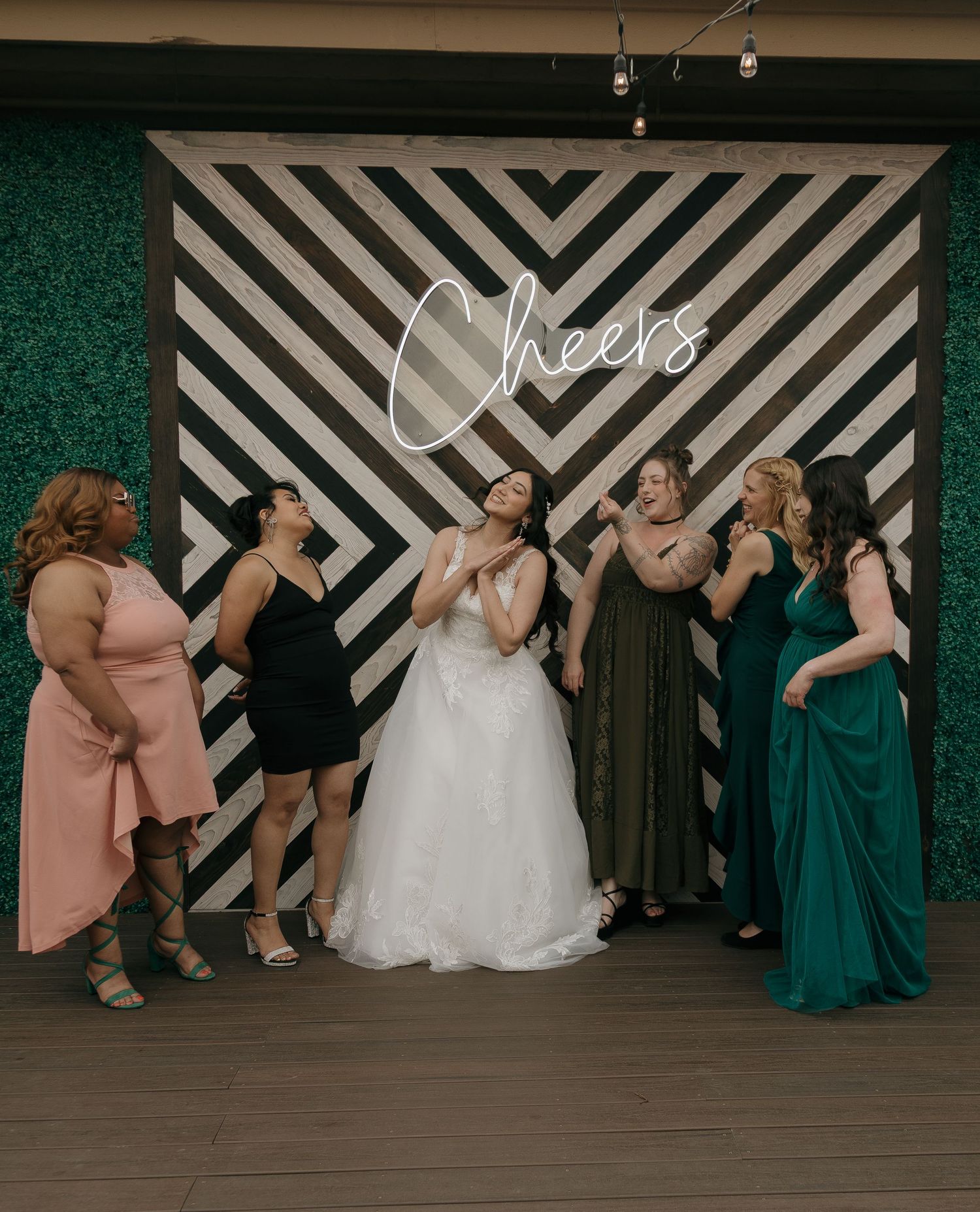 Bride and bridesmaids laugh together in front of a striped Cheers neon sign backdrop.