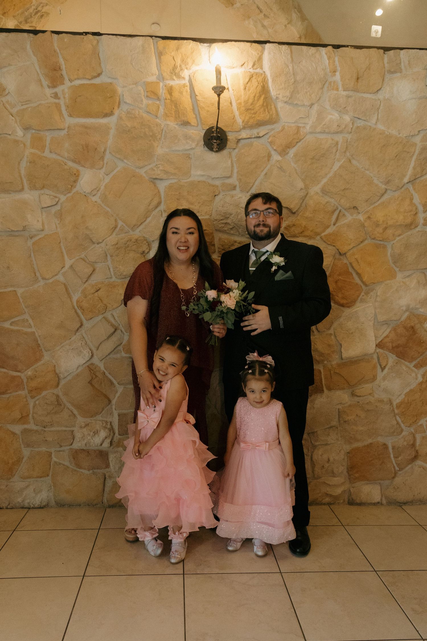 A couple in formal wedding attire poses with two young girls in pink dresses against a stone wall.