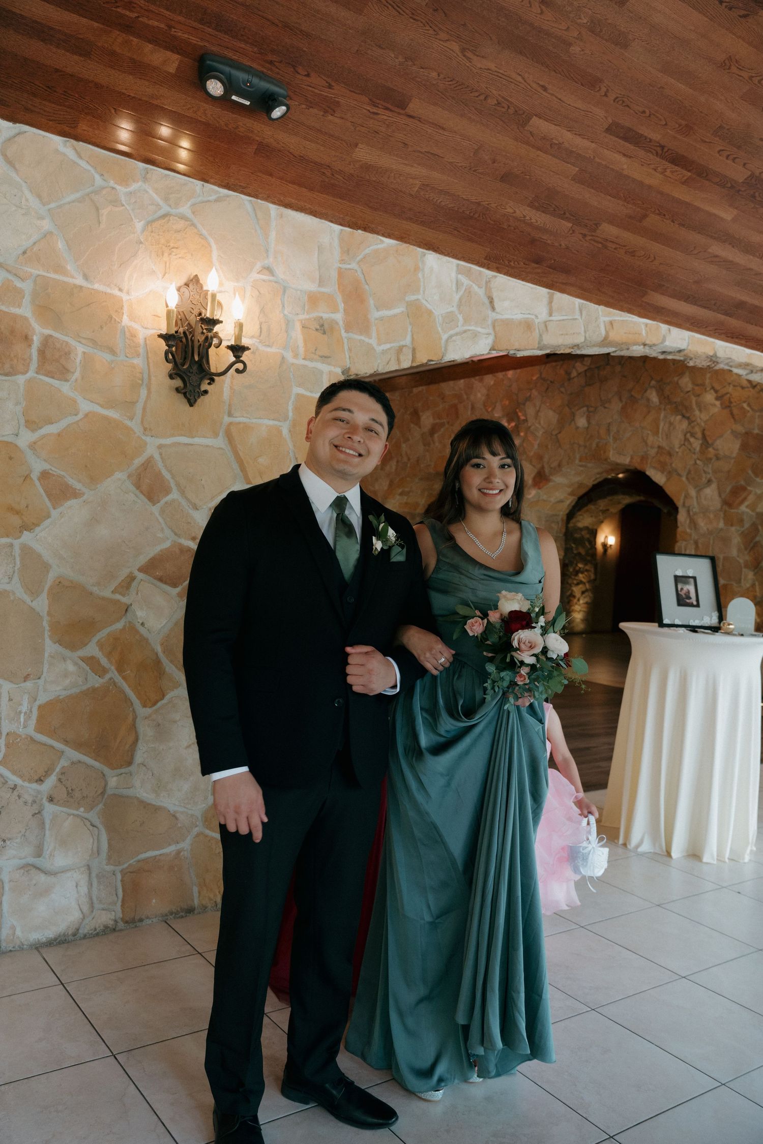 Smiling groom in black suit with green tie poses with bridesmaid in teal gown inside stone-walled wedding venue.