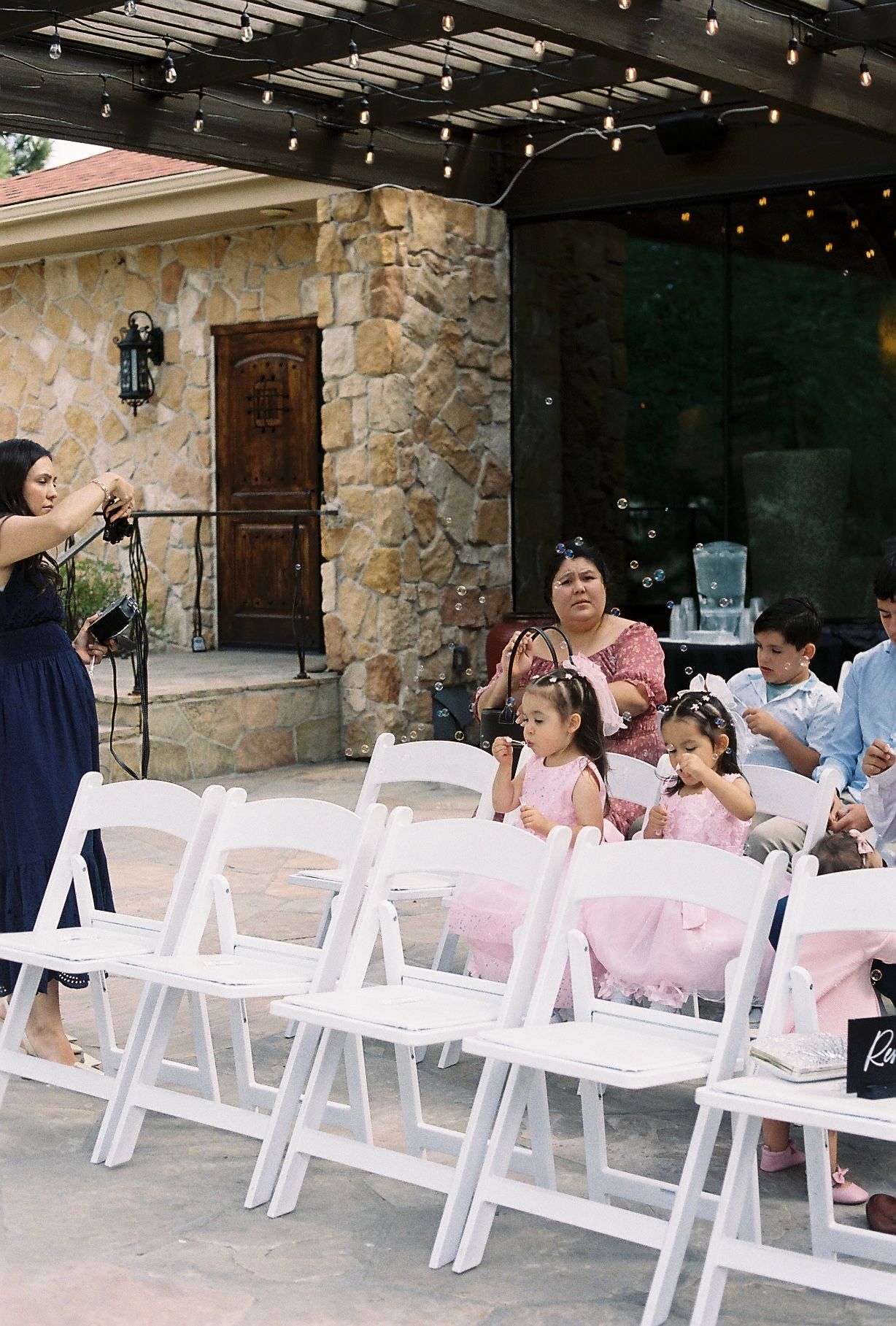 Wedding guests seated in white chairs at an outdoor stone venue ceremony with string lights.