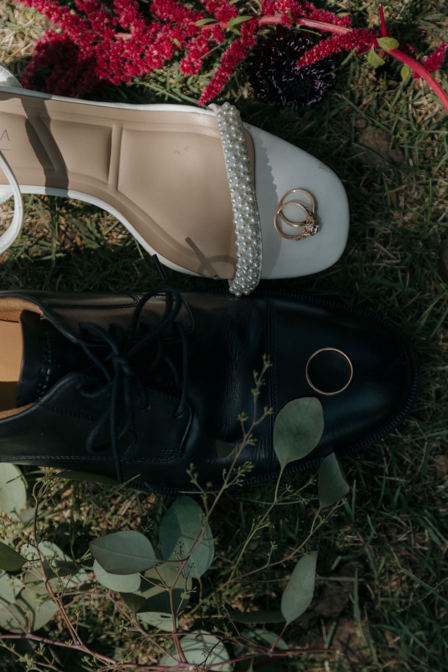 Wedding rings resting on white pearl-adorned bridal heels beside black groom dress shoes on green grass.
