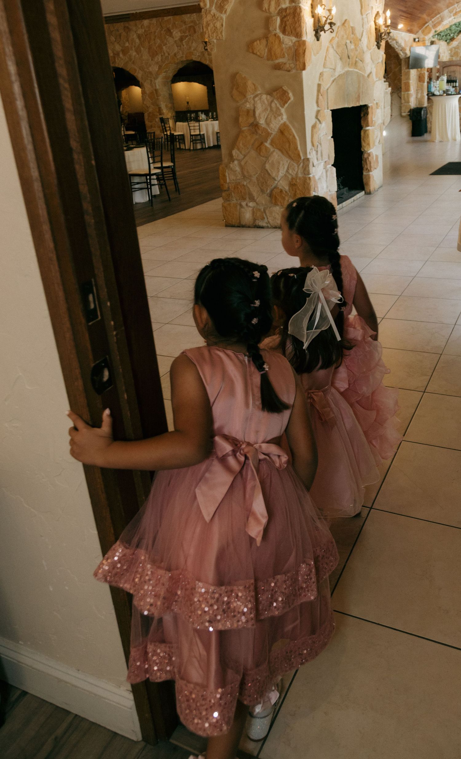 Two flower girls in pink sequin dresses walking through an elegant stone wedding venue corridor.