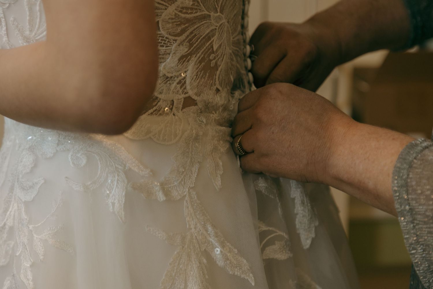 Close-up of hands fastening buttons on intricate lace and feather detail white wedding gown back.
