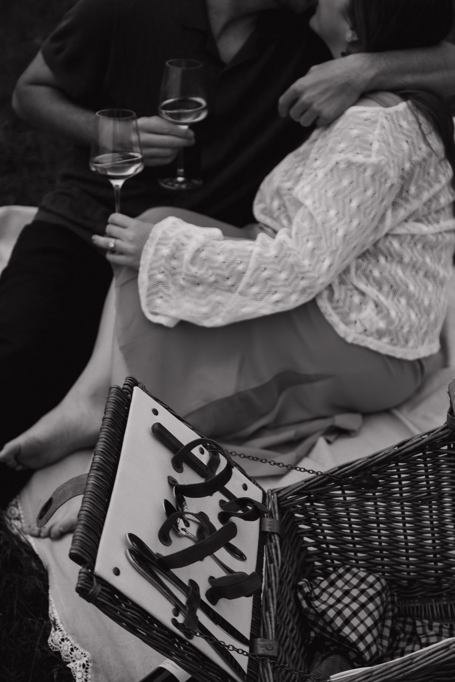 Black and white photo of a couple enjoying a romantic picnic with wine glasses and a wicker basket outdoors.
