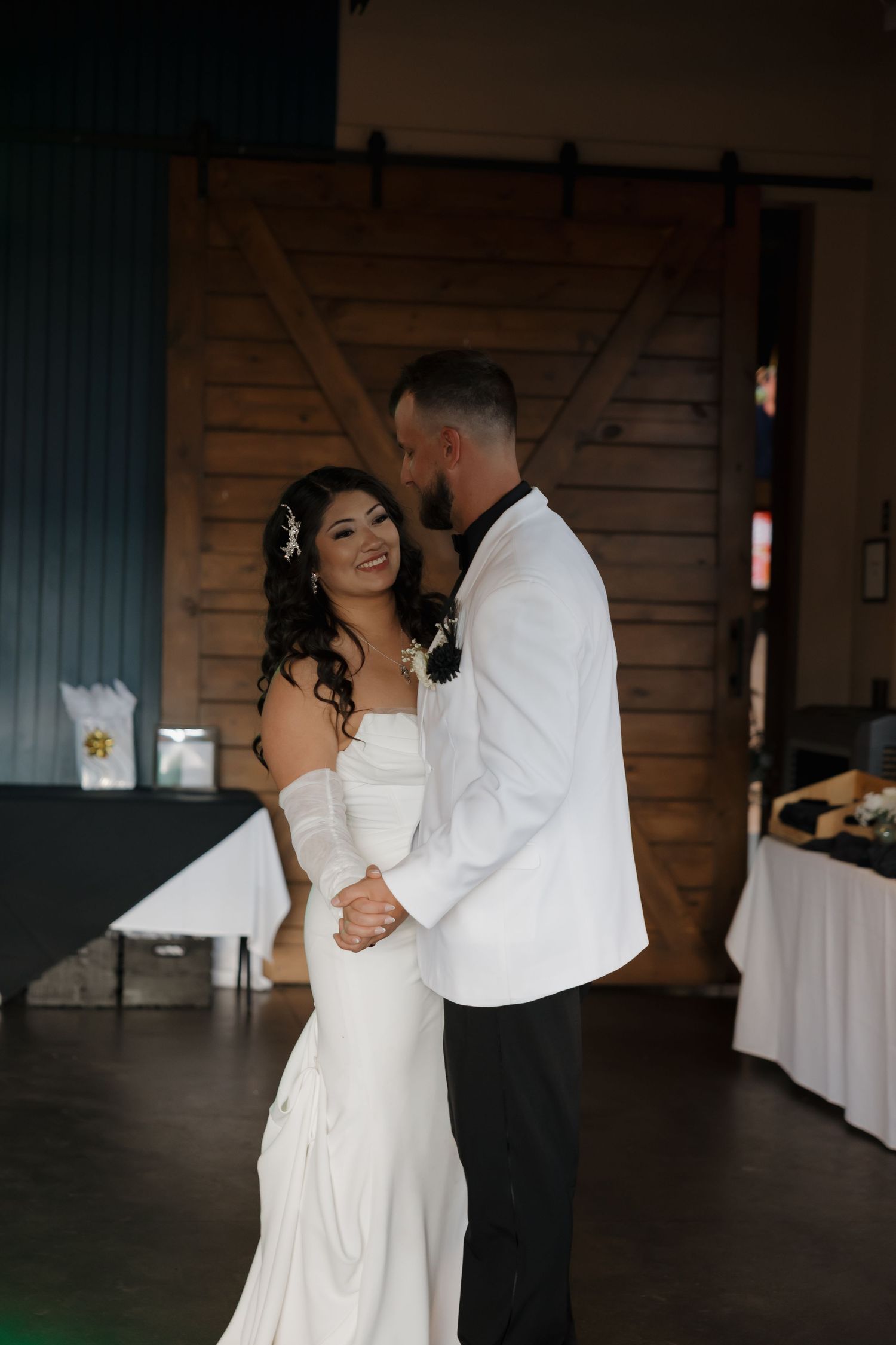 Smiling bride in white gown dancing with groom in white blazer at wedding reception.