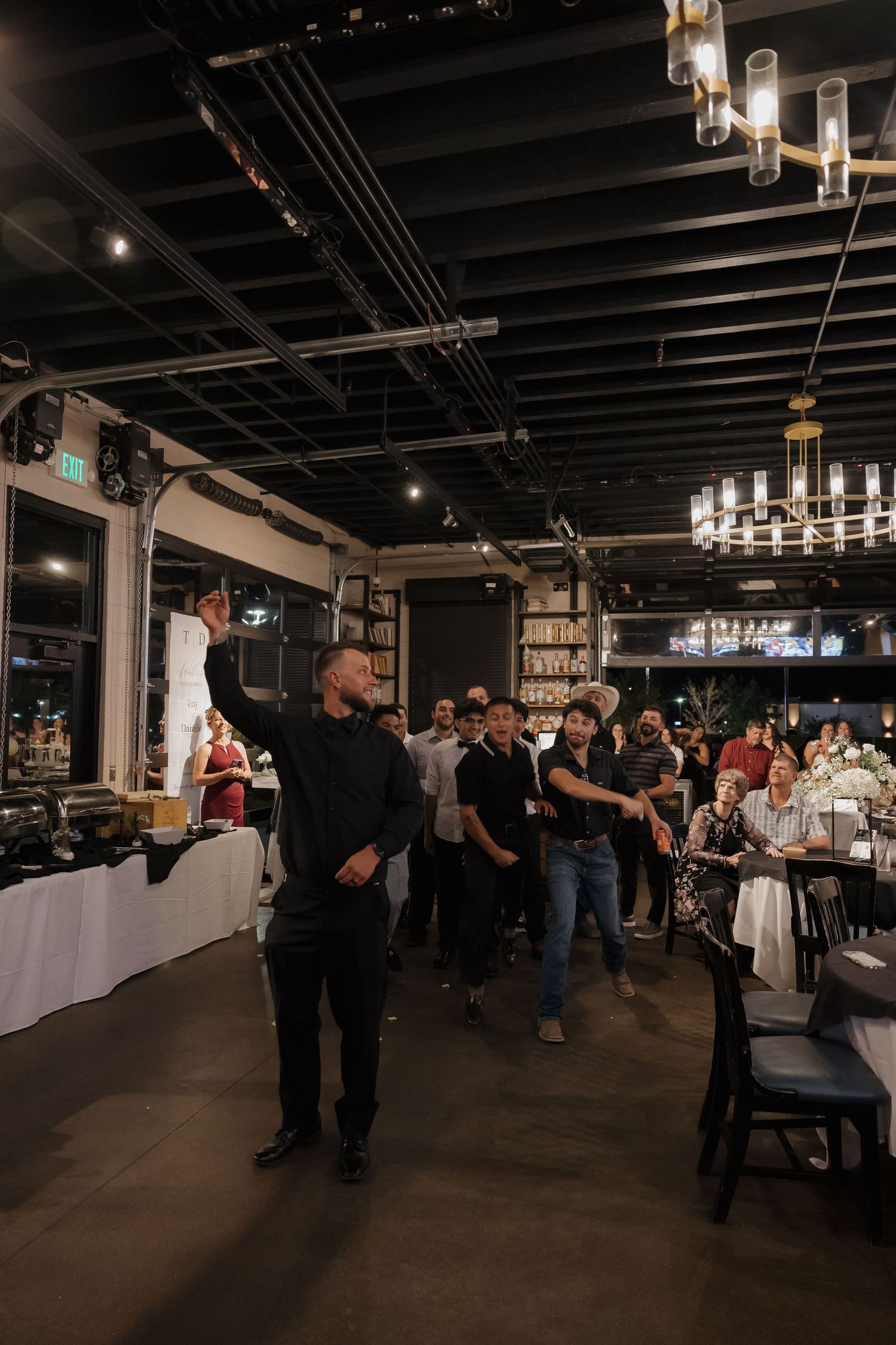 Man in black outfit gestures at an upscale event venue with guests gathered and elegant chandeliers overhead.