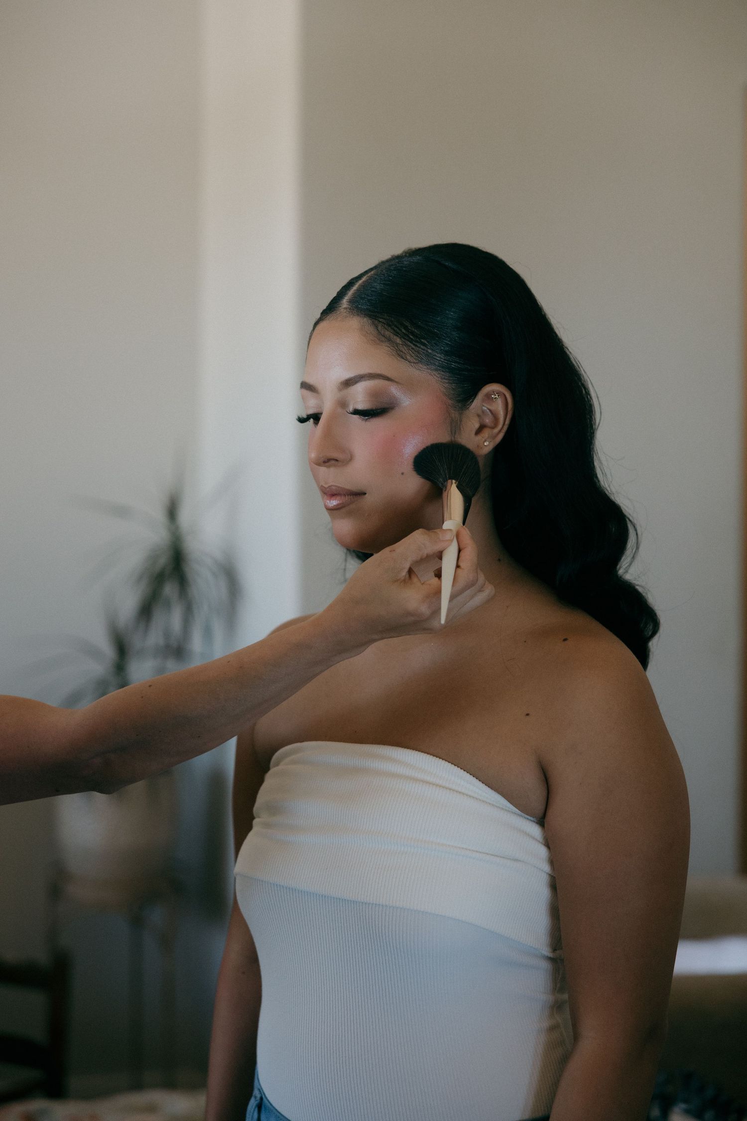 Makeup artist applying blush to a bride's cheek with a large powder brush before her wedding.