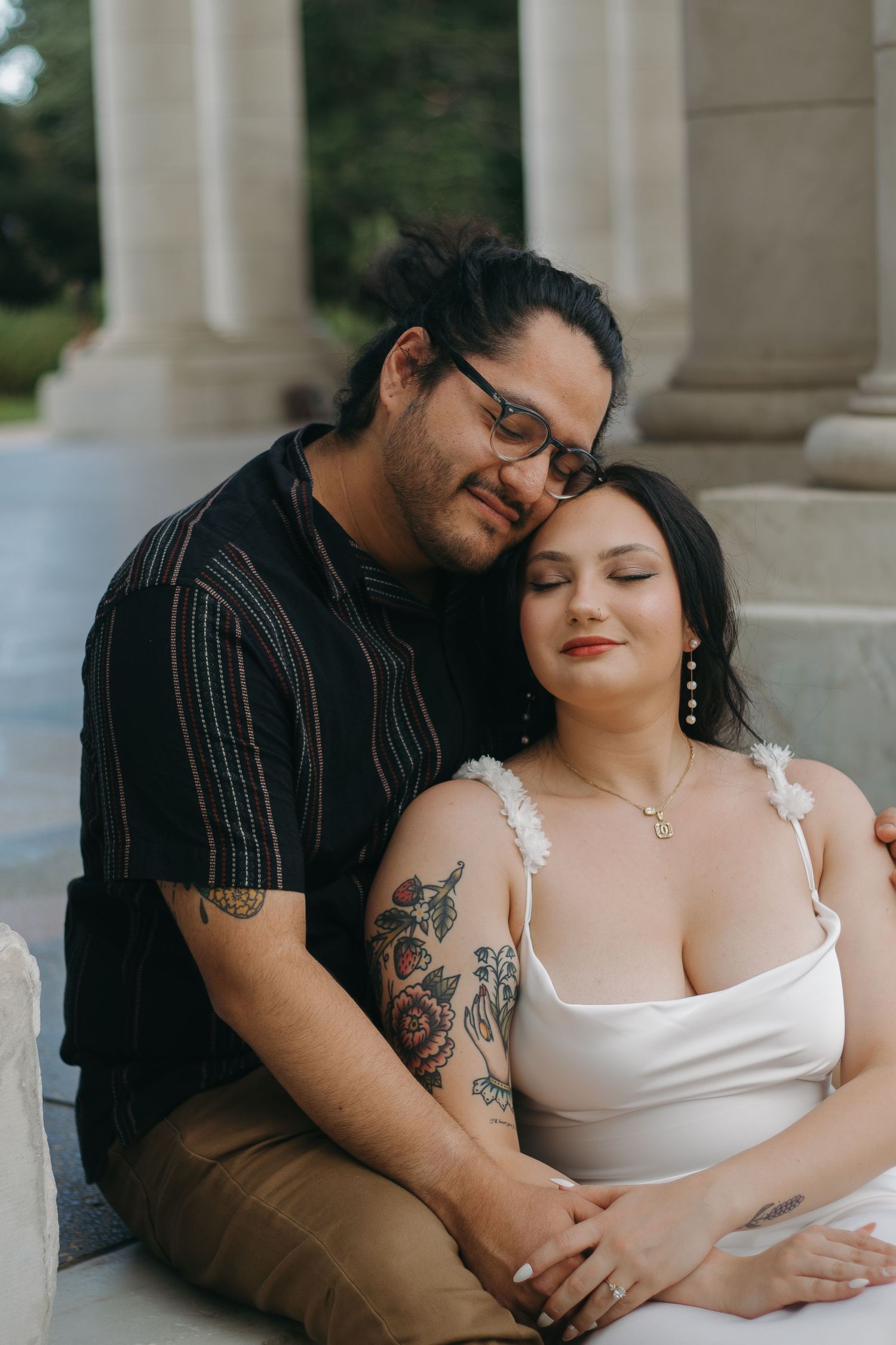 Romantic couple embraces in front of white columns, with one wearing a black striped shirt and the other in white.