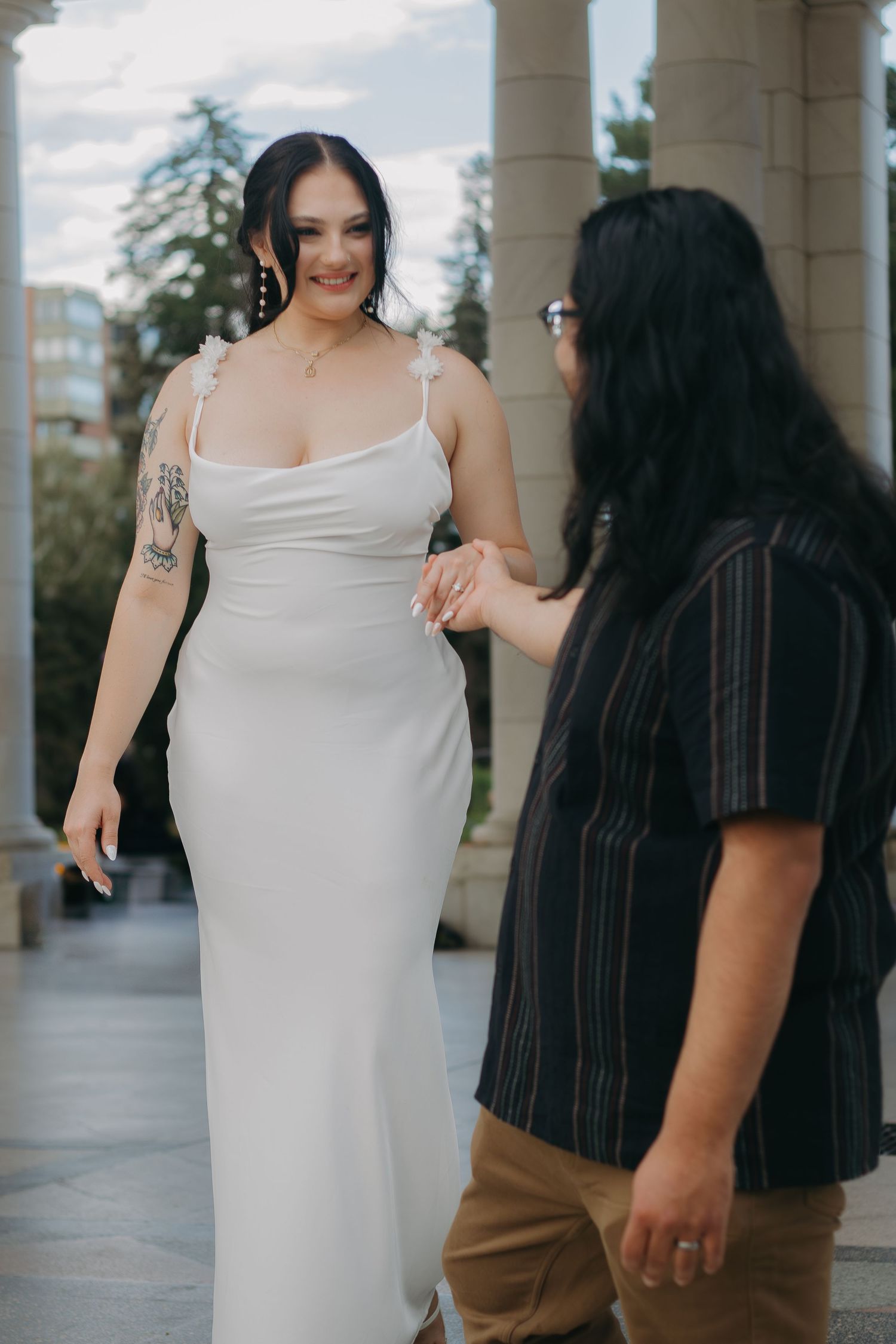 Couple in contrasting outfits stand together, with person in elegant white dress and visible tattoo.