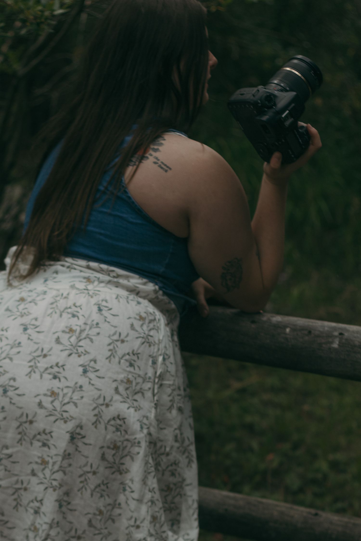 A photographer in a floral dress holds a professional DSLR camera while standing by a wooden railing outdoors.