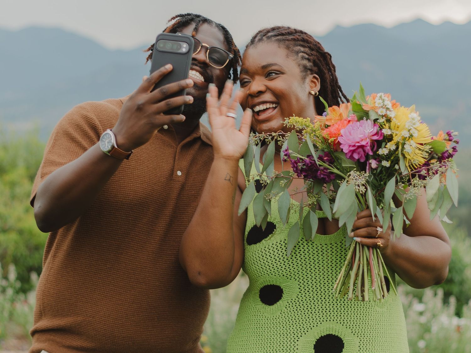 Two people taking a selfie outdoors while one holds a colorful bouquet of flowers on a sunny day with mountains in background.