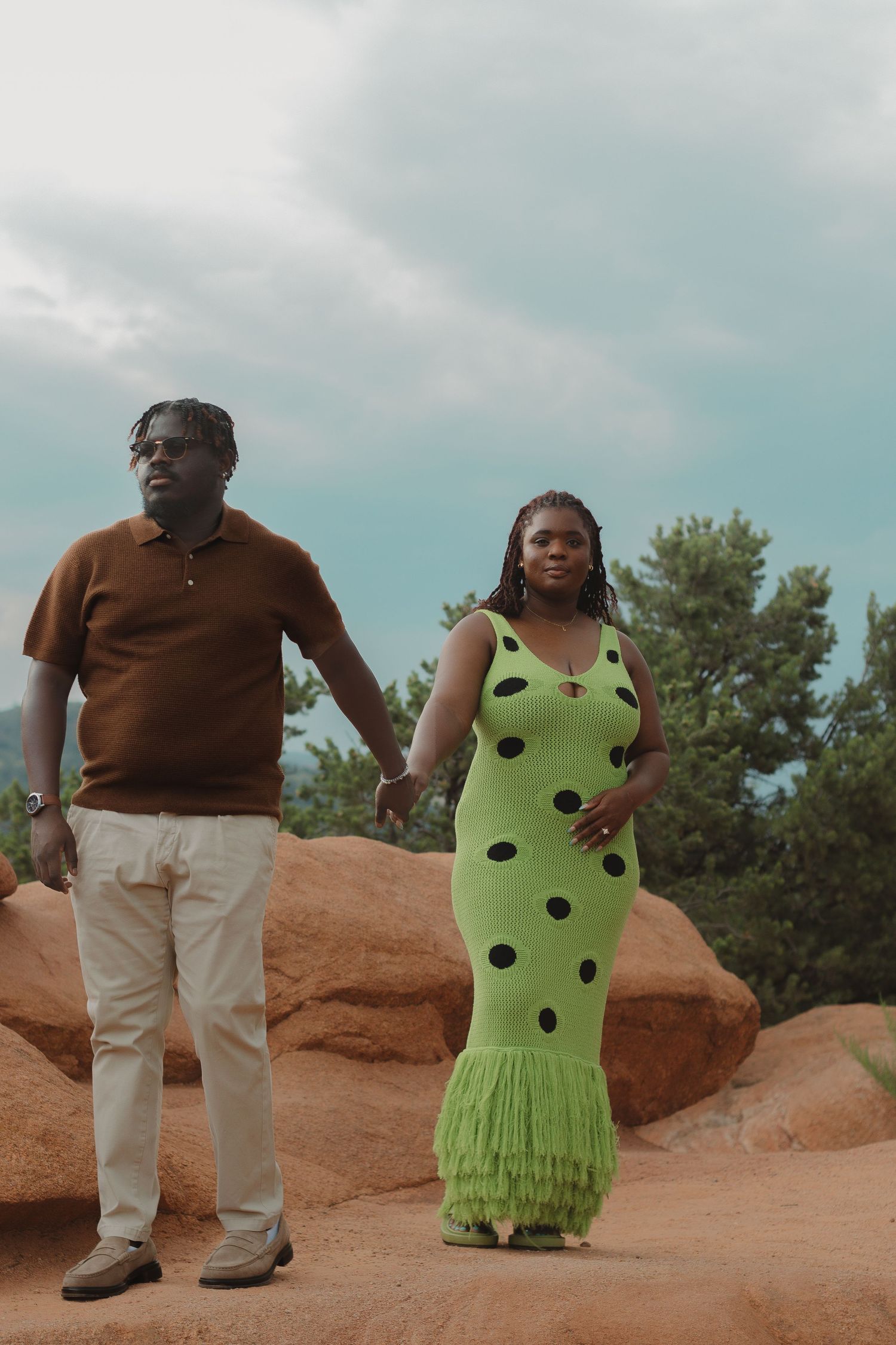 A couple holds hands while standing on red rocks at sunset, wearing brown polo and green polka dot dress with fringe.
