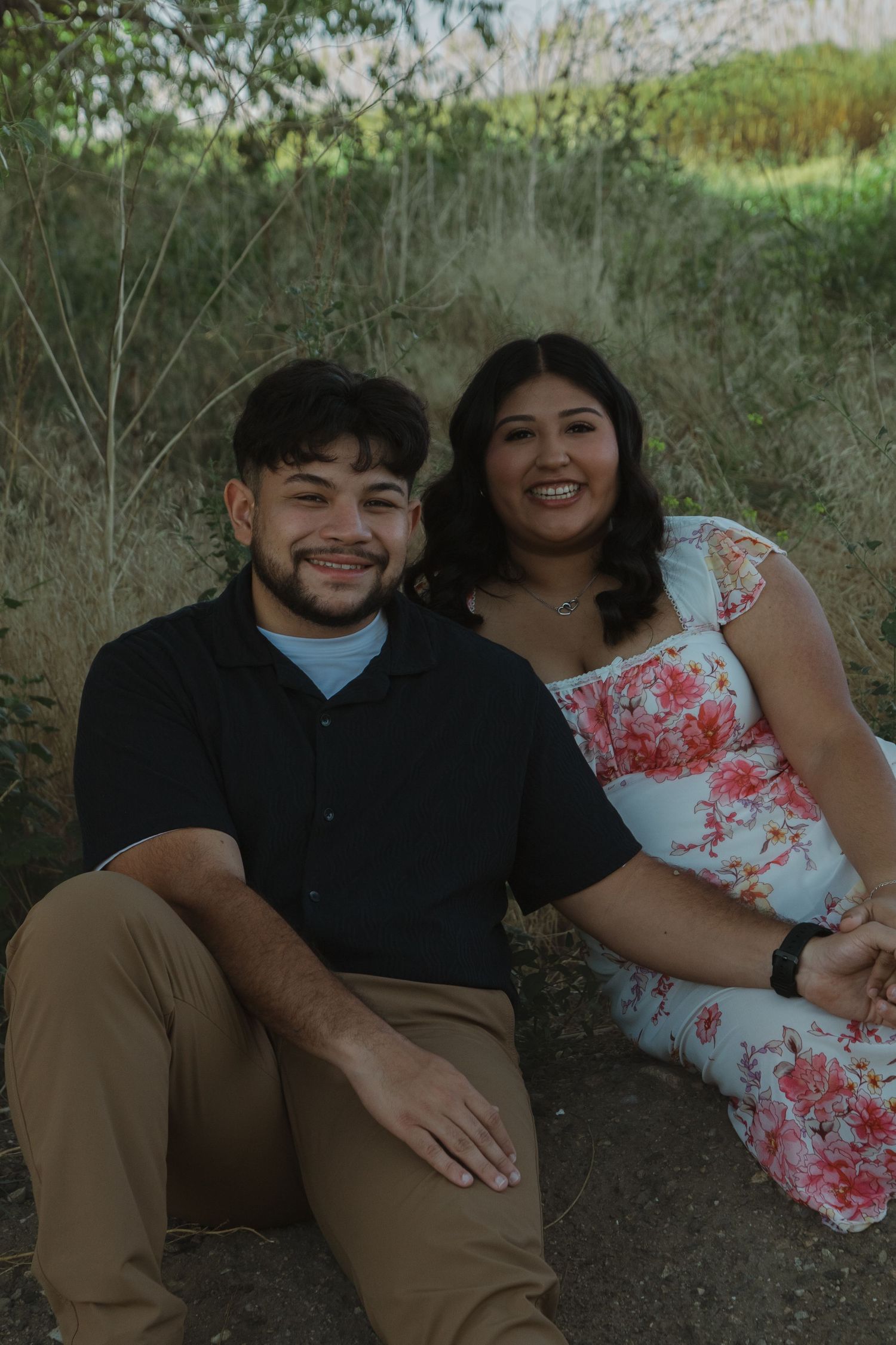 A couple sits together outdoors smiling in casual attire against a natural background.