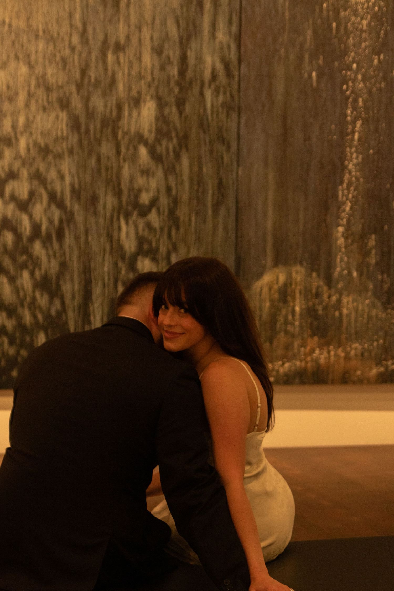 Romantic couple shares intimate moment against textured wall in warm lighting.