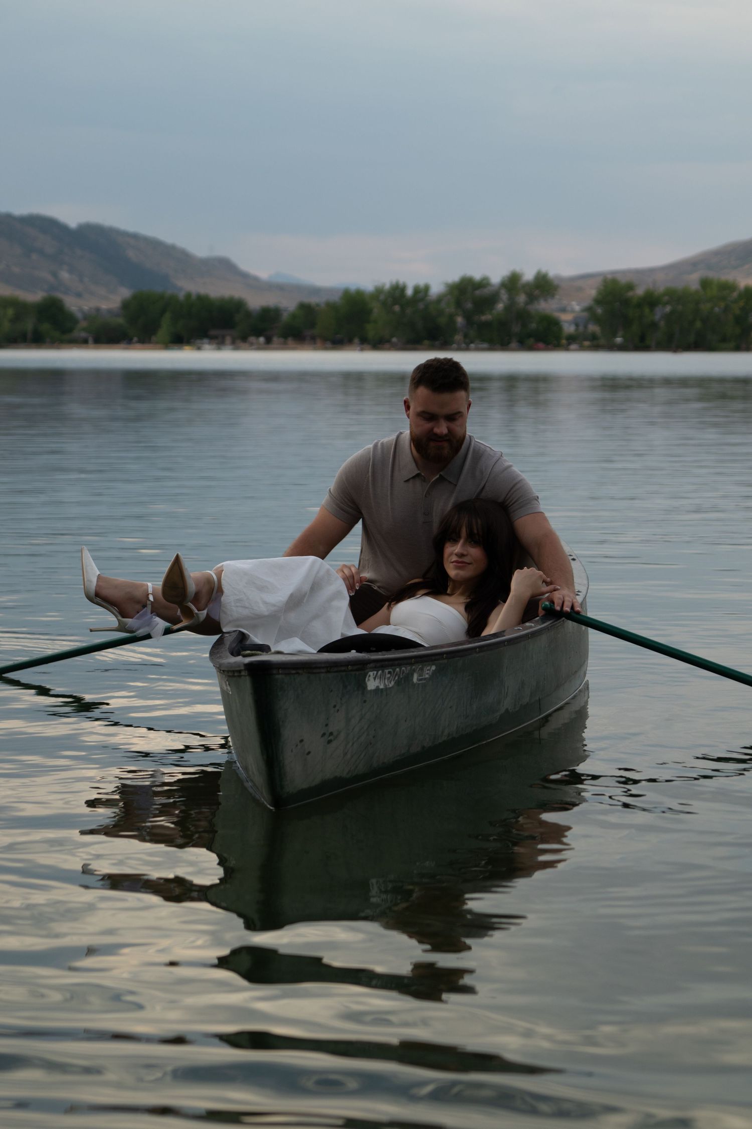 People paddle through calm waters in a dark canoe with mountains and trees visible in the background.