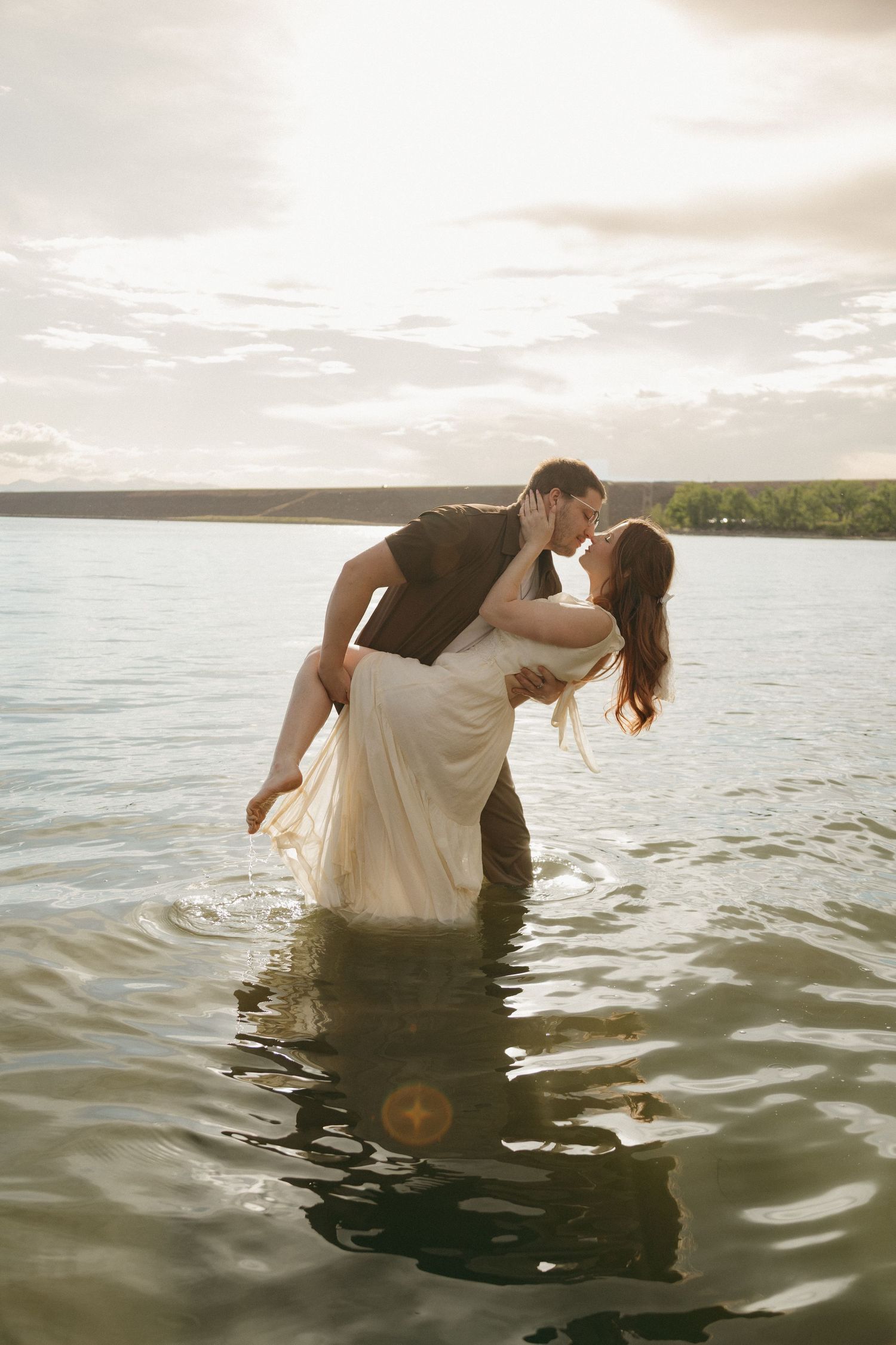 A romantic couple shares a kiss while standing in a lake at sunset, the man carrying the woman in a white dress.