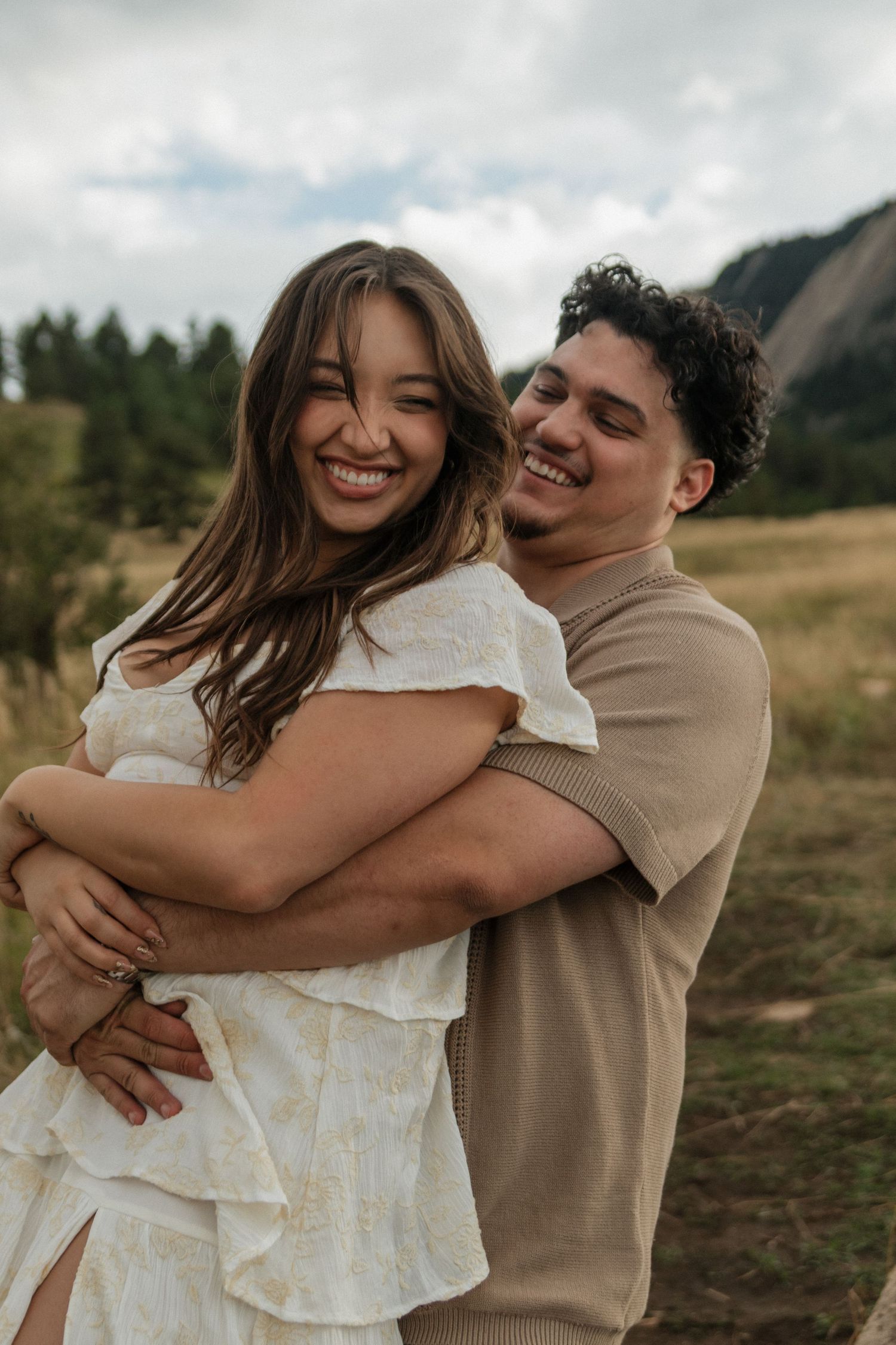 Happy couple laughing and hugging in a mountain meadow, woman in white dress and man in tan shirt.