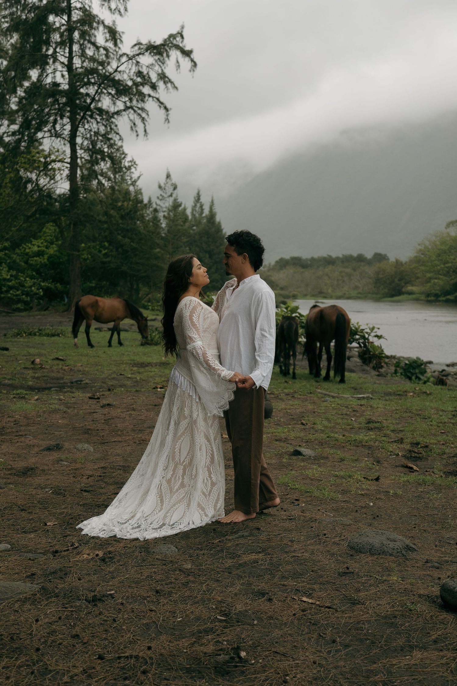 Couple in white lace dress and white shirt hold hands in misty valley with horses grazing by river.