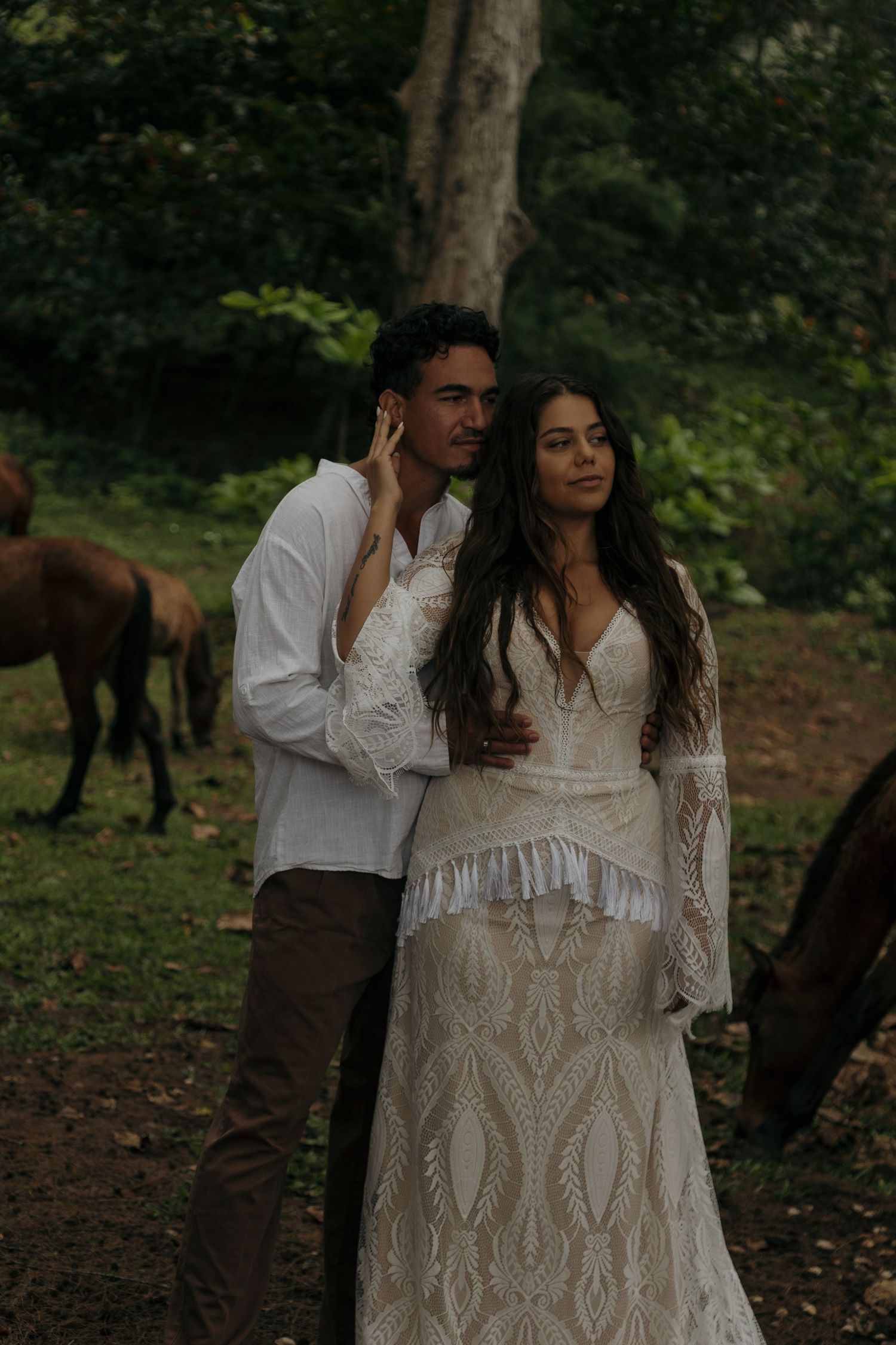 Couple in bohemian wedding attire poses romantically among horses in a lush forest setting.