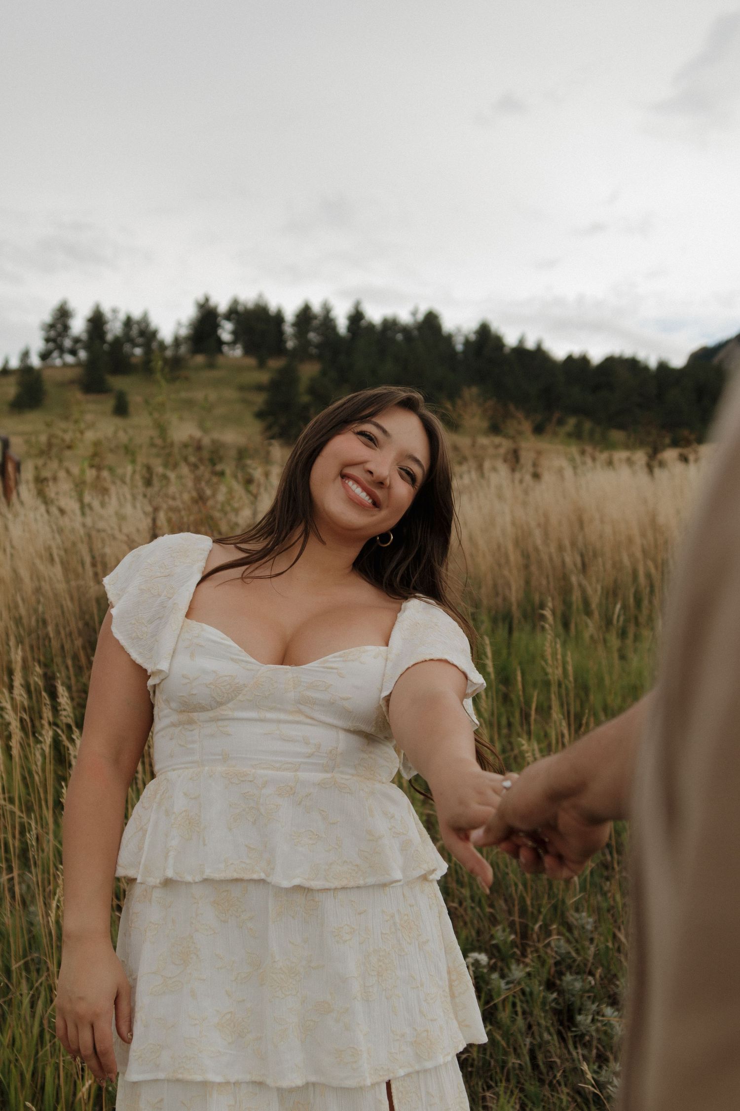 Smiling woman in white floral dress holds hands with partner in golden grass field with pine trees.