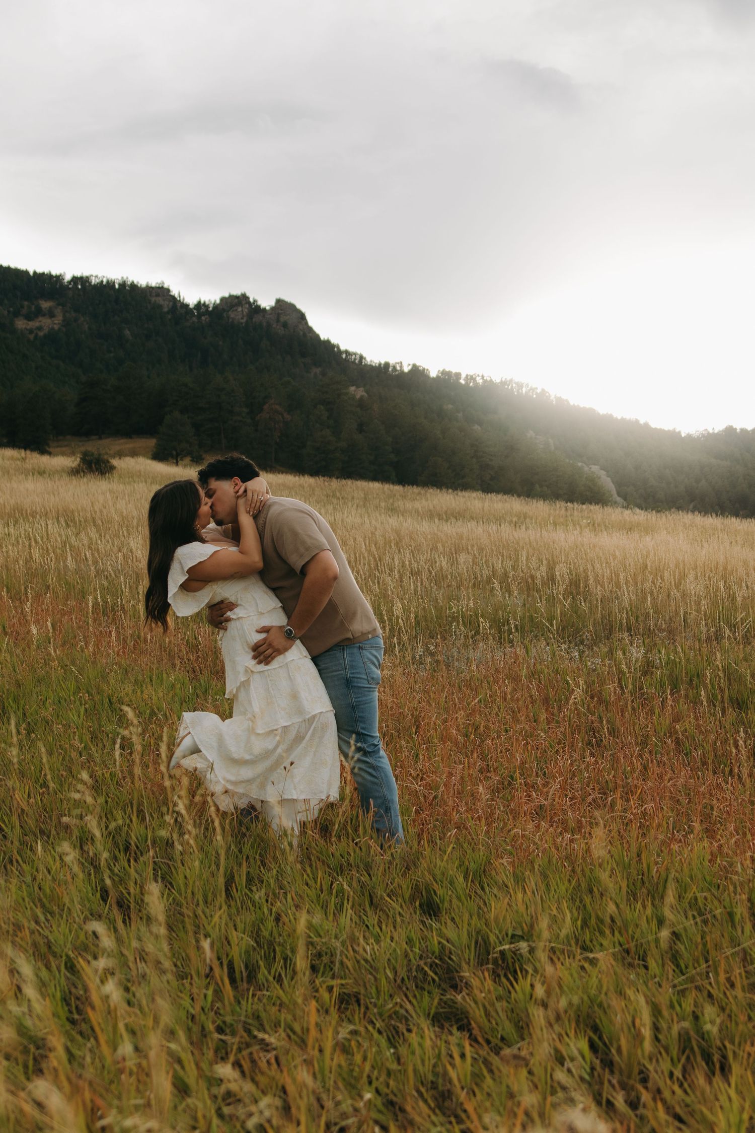 Romantic couple sharing a kiss in a sunlit grassy field with scenic mountain landscape behind them.
