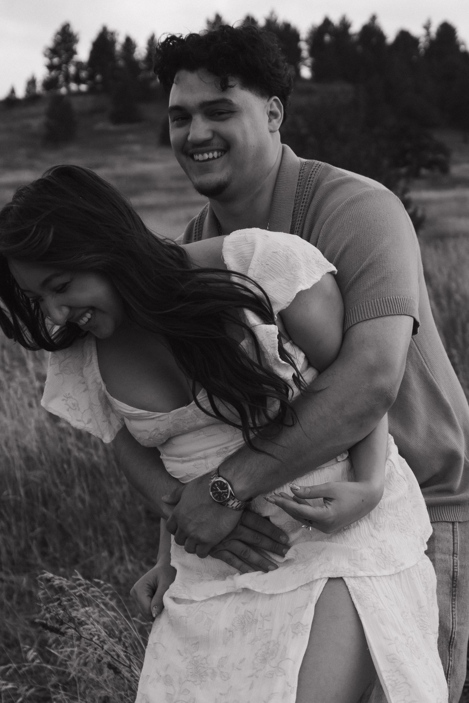 Black and white photo of man hugging laughing woman from behind in grassy outdoor setting.
